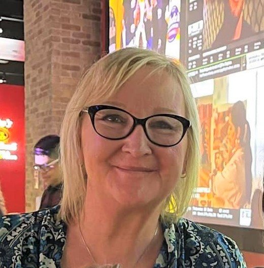 A woman with blonde hair and glasses smiling in a room with animated posters on the wall.