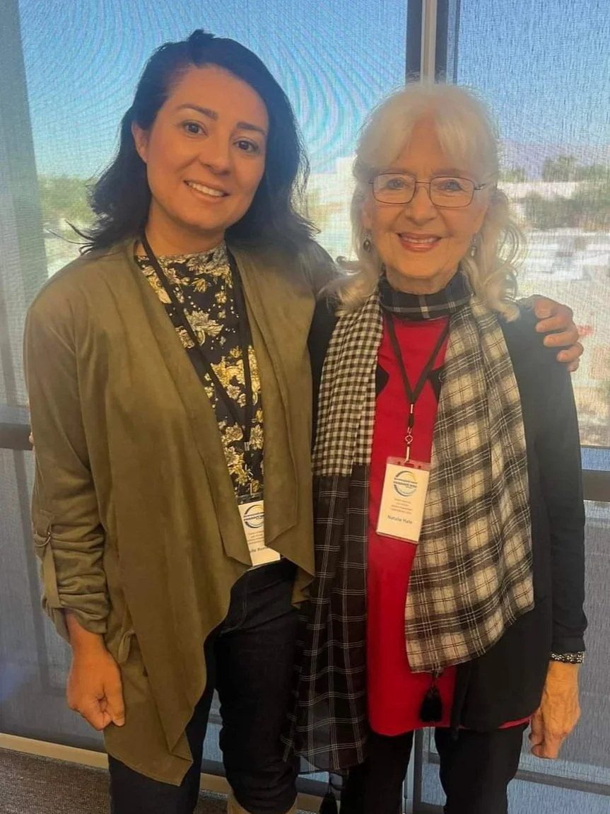 Two women standing together in front of a window, smiling for the camera. They are wearing conference badges around their necks.