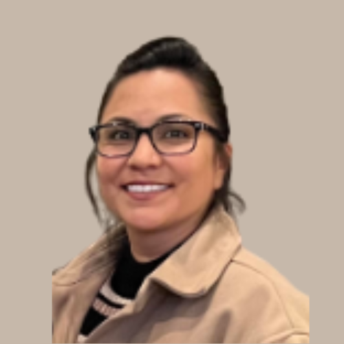 A woman with glasses smiling, wearing a tan jacket and a black shirt with a white stripe, standing against a plain background.