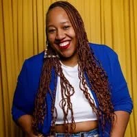A woman with styled dreadlocks, wearing a white T-shirt with purple and gold lettering, smiling and sitting at a wooden table with a blue background.