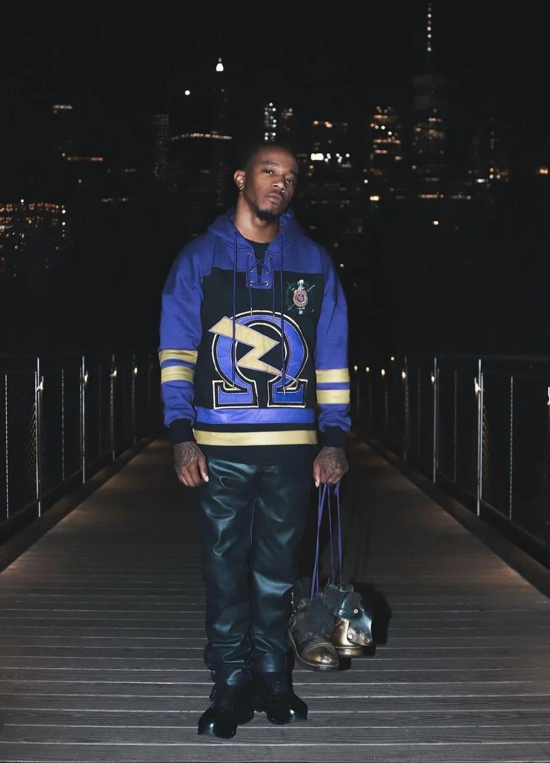 A man standing on a wooden pier at night, holding a pair of black and gold shoes, with a city skyline lit up in the background. He is wearing a blue and black athletic hoodie with yellow accents, black pants, and shiny black shoes.