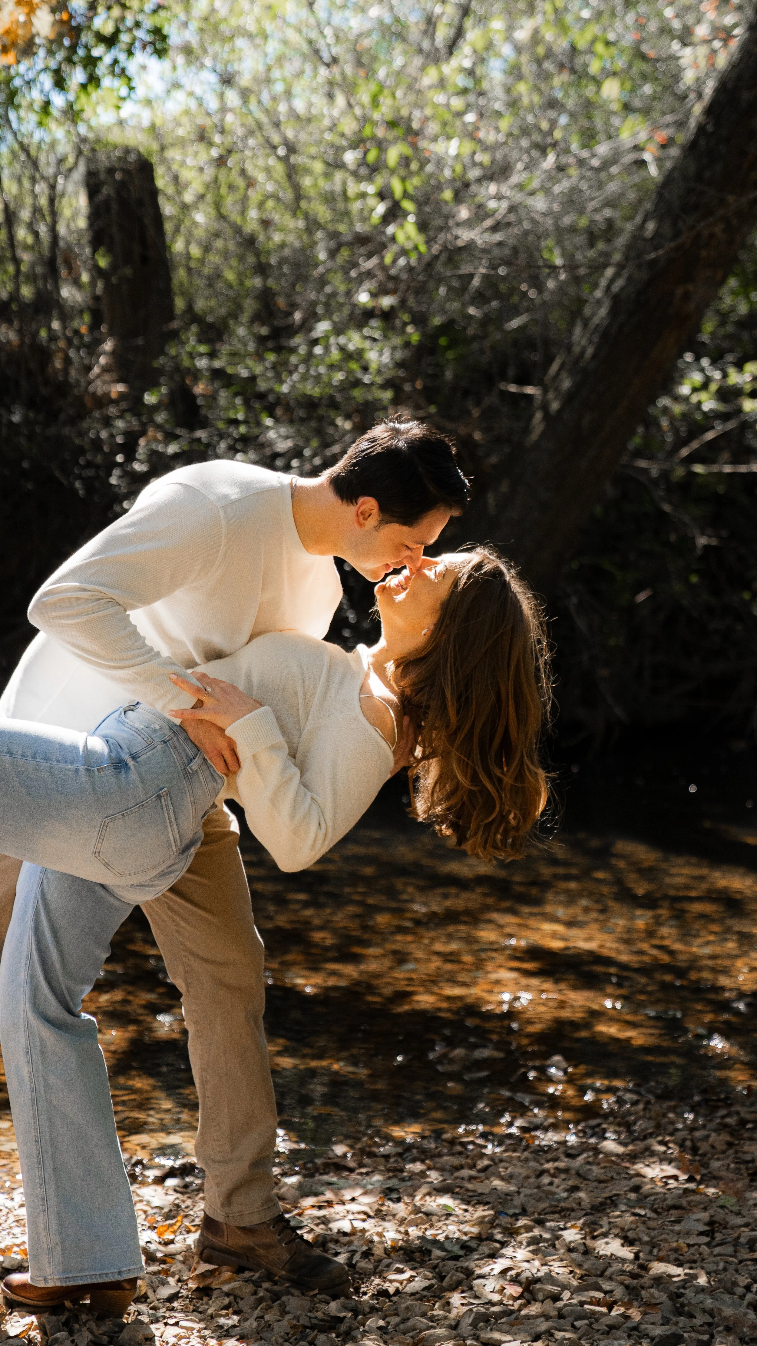 A couple is dancing and smiling on a rocky riverbank surrounded by trees.