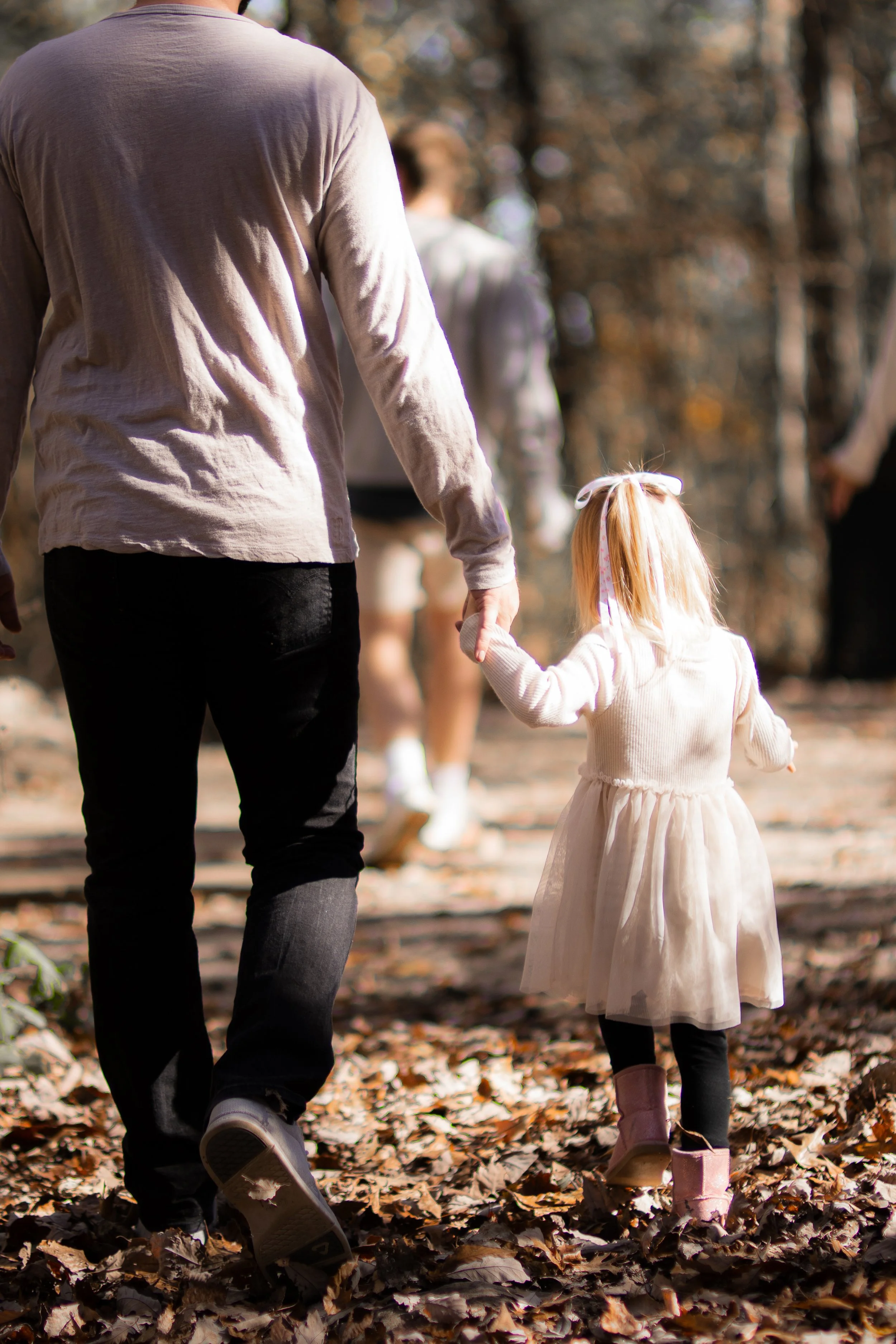 A man and a young girl holding hands while walking in a forest during autumn, with fallen leaves on the ground.