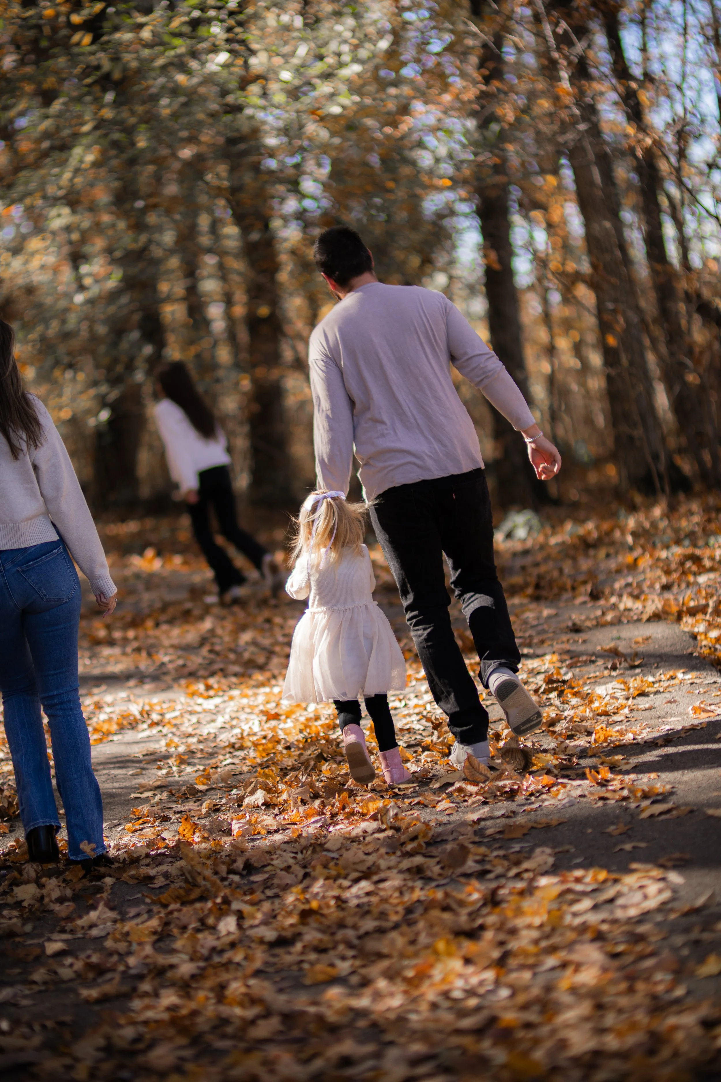 A man and a little girl walk on a leaf-covered trail in a forest during autumn, with other people visible in the background.