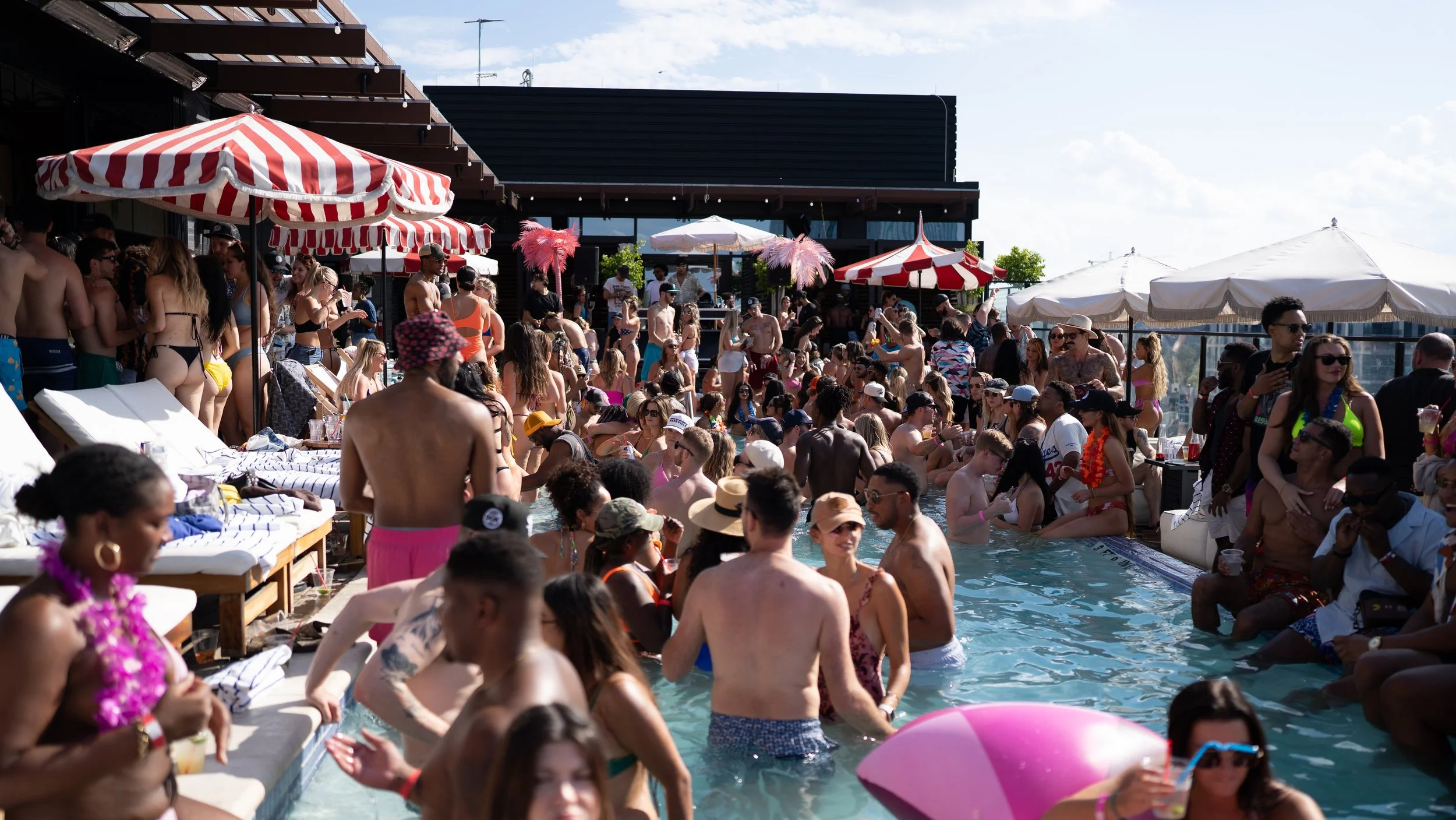 Crowd of people at a rooftop pool party with umbrellas, sunbathers, and some dancing under a partly cloudy sky.