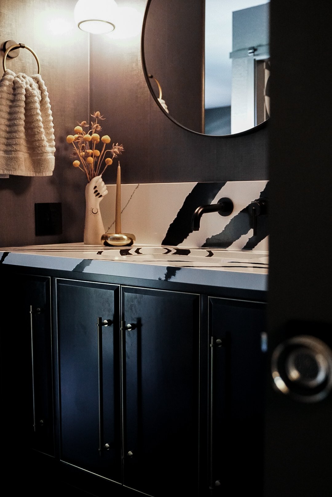 Modern bathroom vanity with black cabinets, a black and white patterned countertop, a round mirror, a decorative hand-shaped vase with dried flowers, and a beige towel hanging on a towel ring.