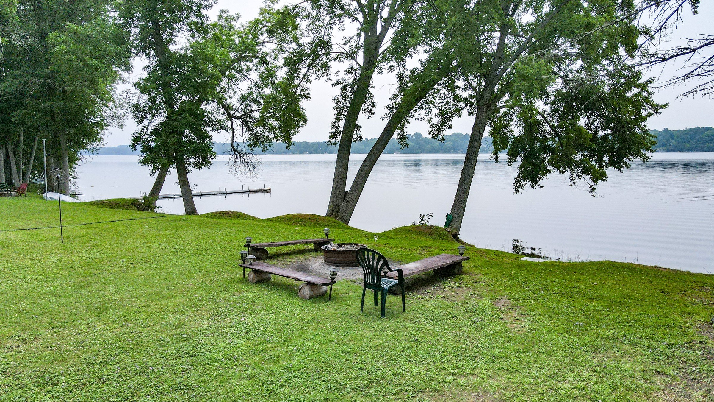 Lakeside firepit ringed by log benches on a grassy shoreline at Lake Dalrymple, with a dock reaching into the water beyond, Ontario