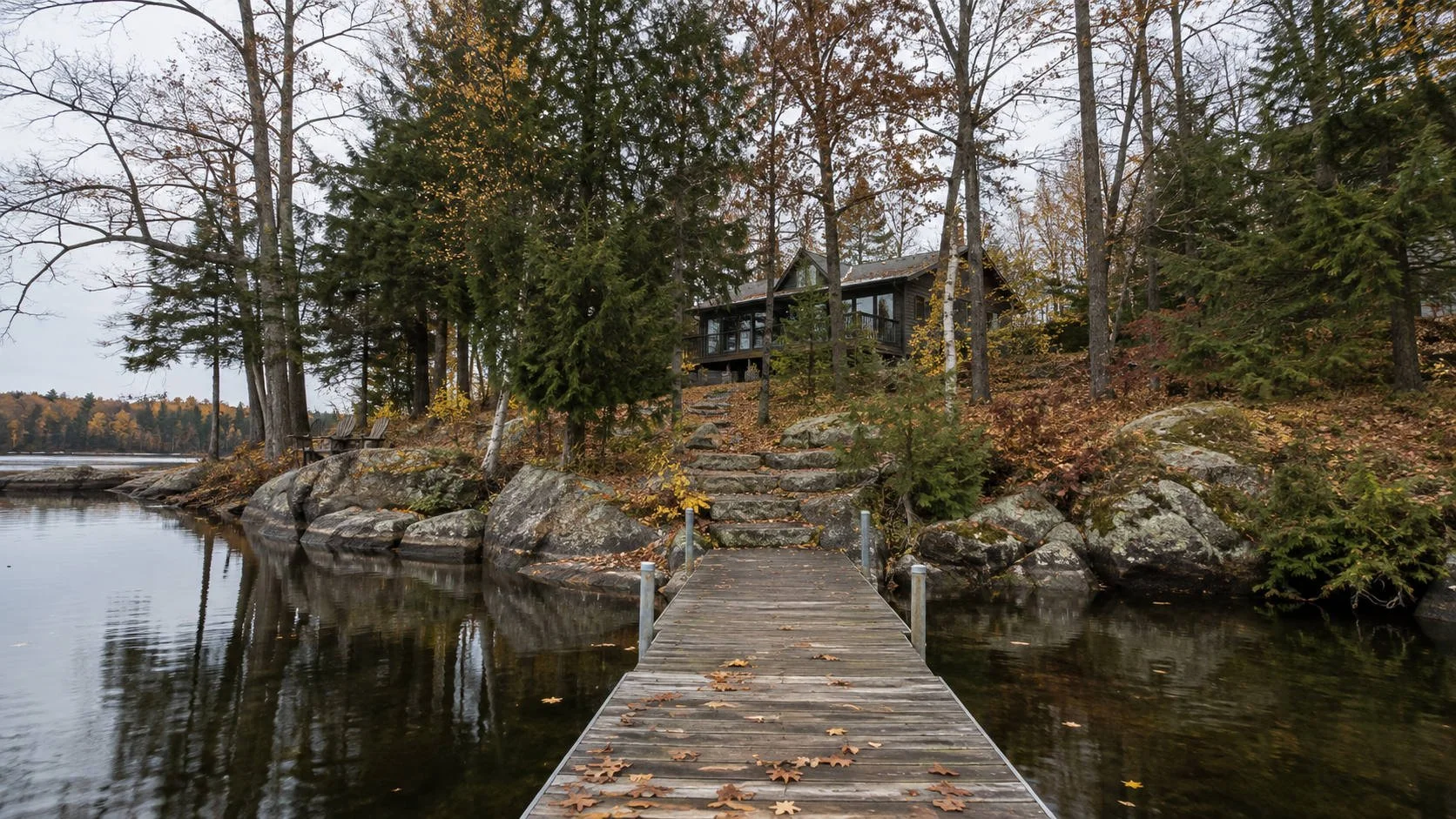 A wooden dock scattered with fallen leaves looking back at a cottage on a rocky Ontario shoreline in late autumn