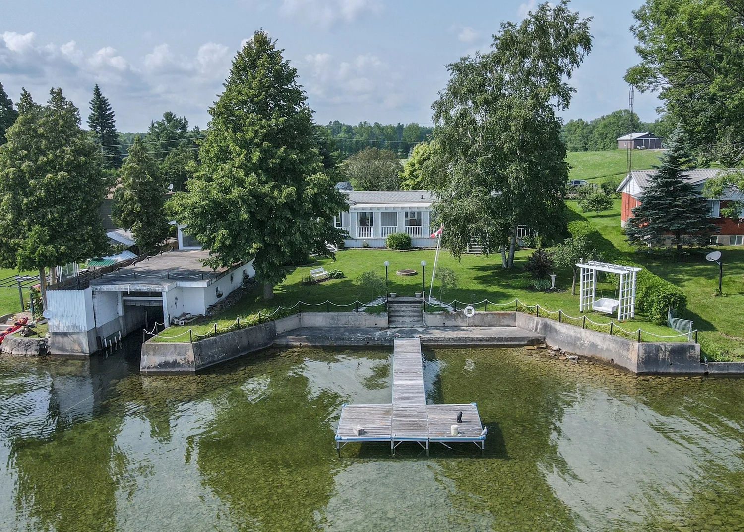 Aerial view of a Lake Simcoe waterfront cottage with a T-shaped dock, concrete shoreline wall and clear shallow water on Barnstable Bay