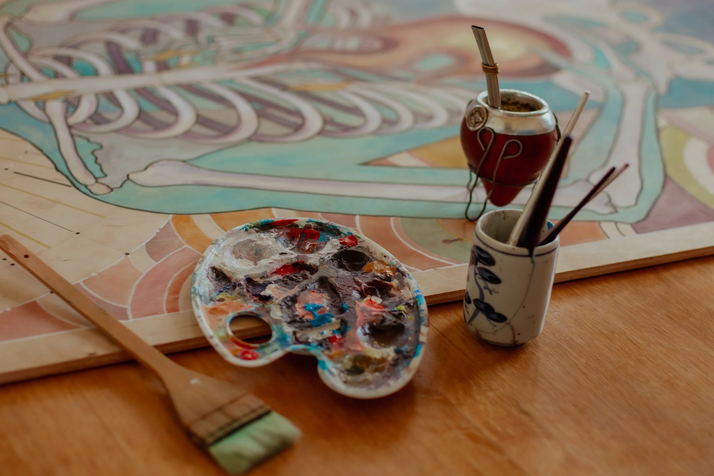 Art supplies including a paint palette with mixed colors, a paintbrush, and a small cup with brushes on a wooden table, with a colorful drawing of a human skeleton on paper in the background.