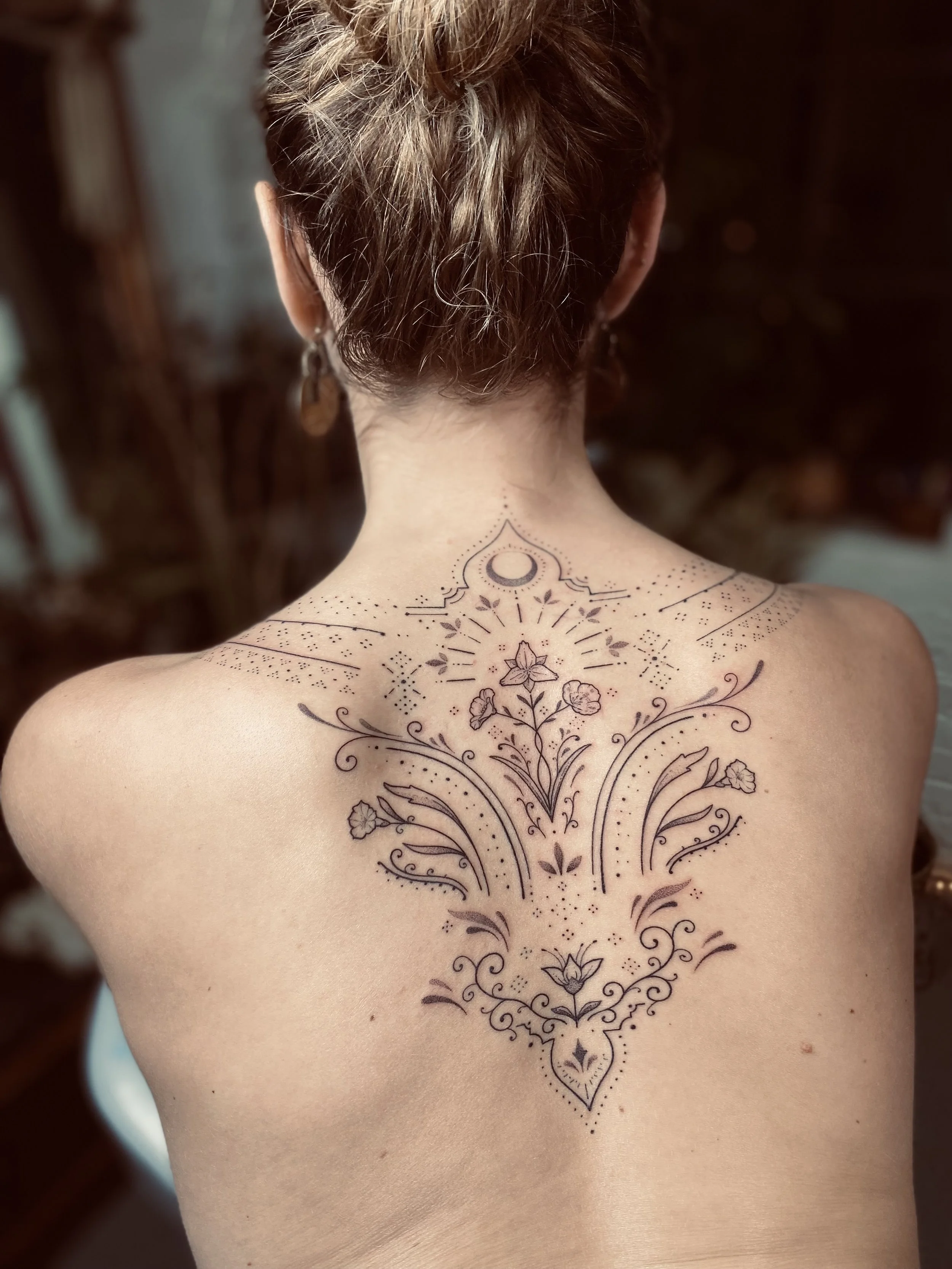 Back view of a woman with a detailed floral and geometric tattoo on her upper back and neck.