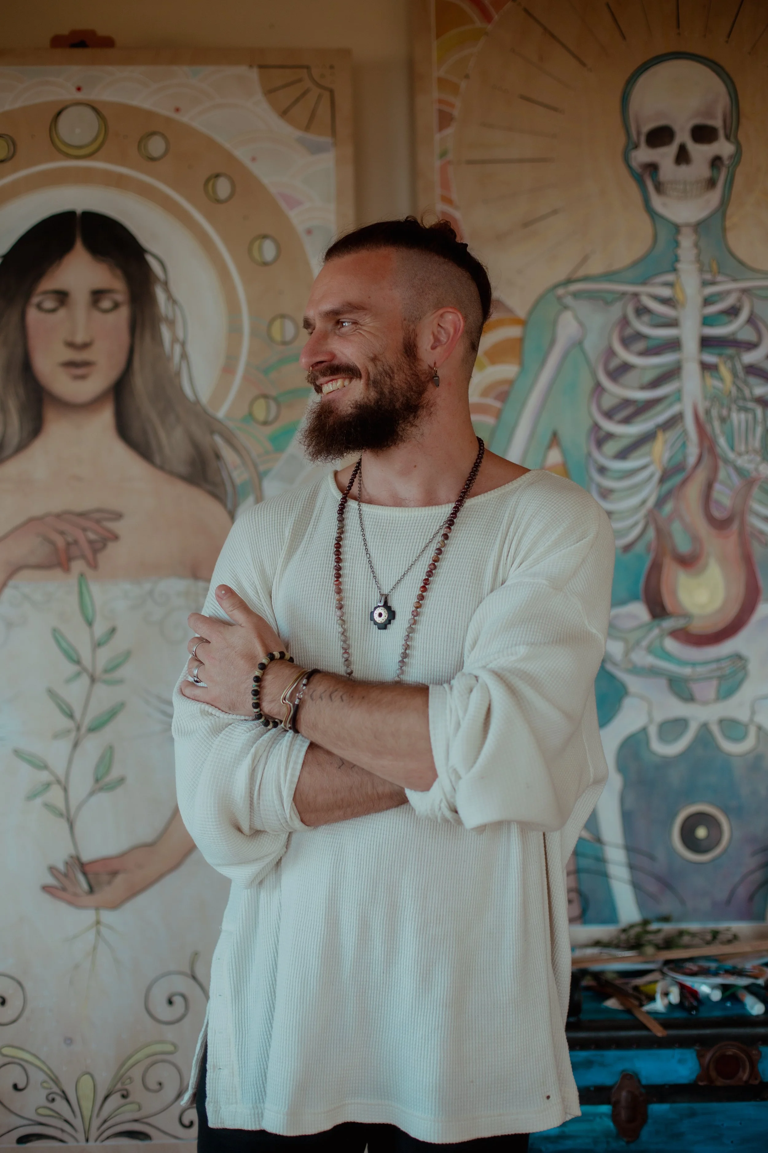 A man with a beard, earrings, and necklaces wearing a white long-sleeve shirt, standing with arms crossed in front of colorful murals of a woman and a skeleton on the wall behind him.