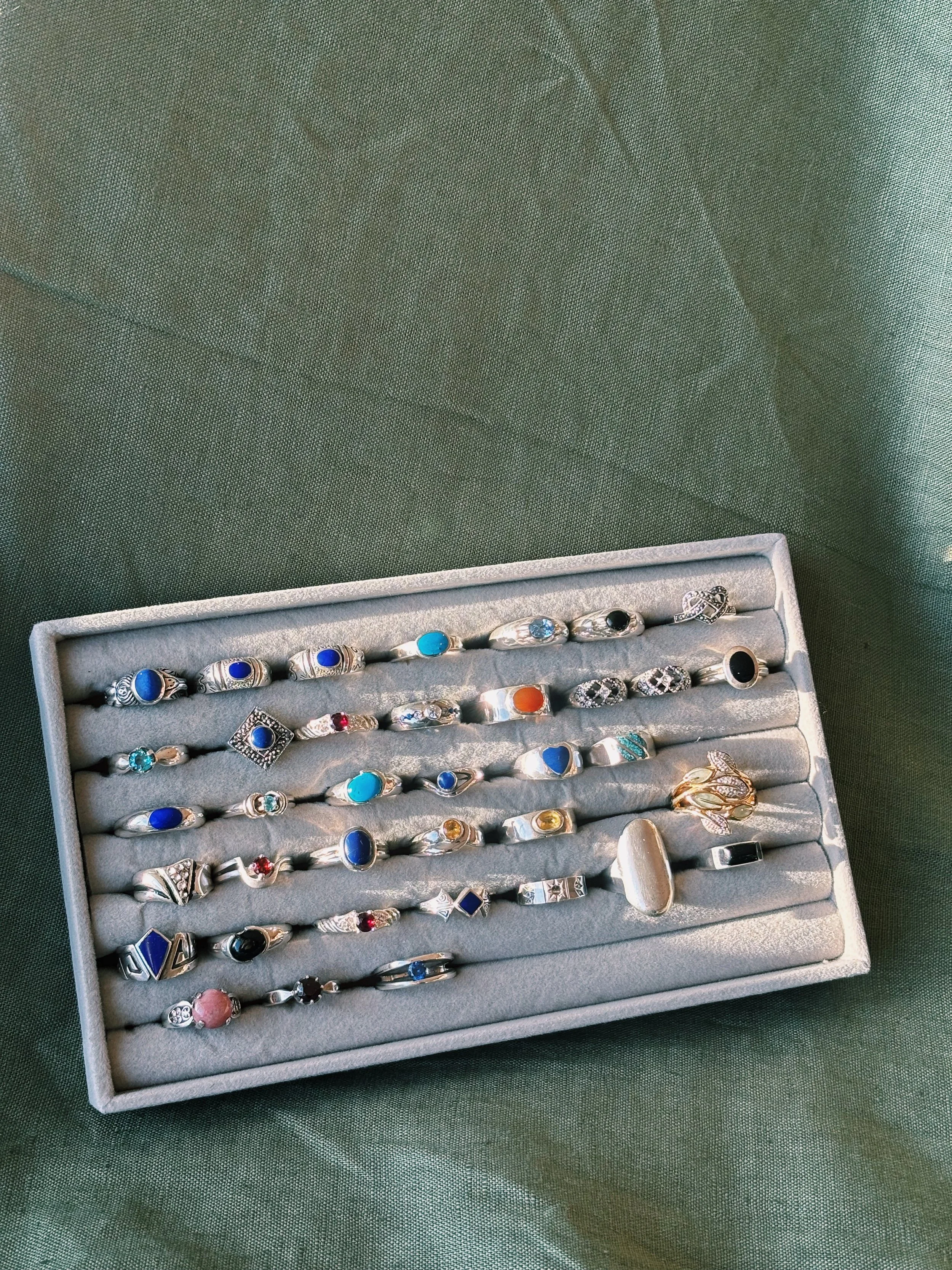 A jewelry box displaying multiple silver rings with various blue, black, red, pink, and clear gemstones, arranged in rows on a grey padded interior.