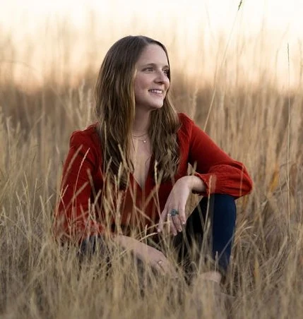 Young blonde woman sitting in a field of tall grass, smiling and looking into the distance, wearing a red top.
