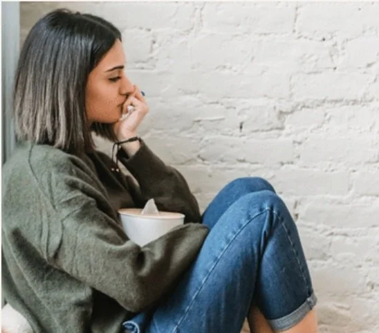 A young brunette woman sitting against a white brick wall, holding a bowl, and resting her head on her hand, appearing pensive.