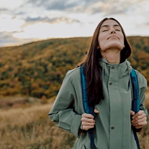 Woman with closed eyes and a peaceful expression, wearing a green jacket and a blue backpack, outdoors in a scenic landscape with hills and cloudy sky.