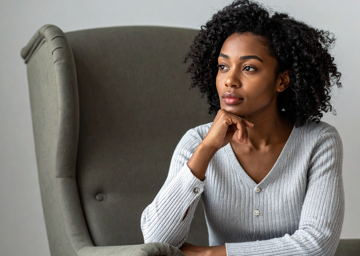 A woman with curly black hair sitting in a green armchair, wearing a gray ribbed cardigan, looking contemplative with her chin resting on her hand.