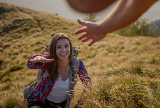 A woman with a backpack smiling as she climbs a grassy hillside, reaching out her hand toward a person above her.