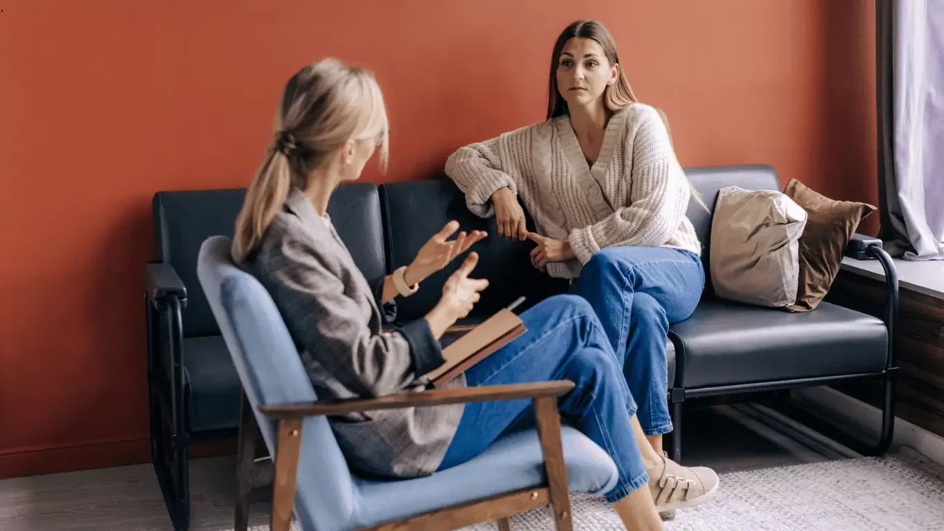 Two women sitting on a black couch in a therapy session. One woman, with blonde hair, is talking and holding a book. The other woman, with brown hair, is listening attentively, leaning on the armrest of the couch.