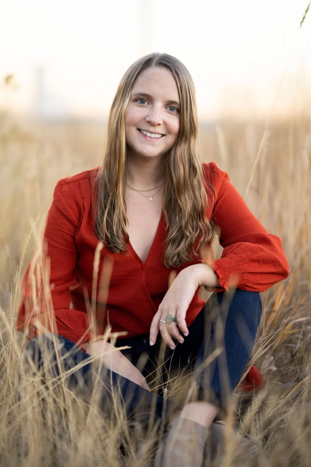 Young woman with long brown hair smiling, sitting in a field of tall, golden grass, wearing a red long-sleeve shirt and blue jeans.