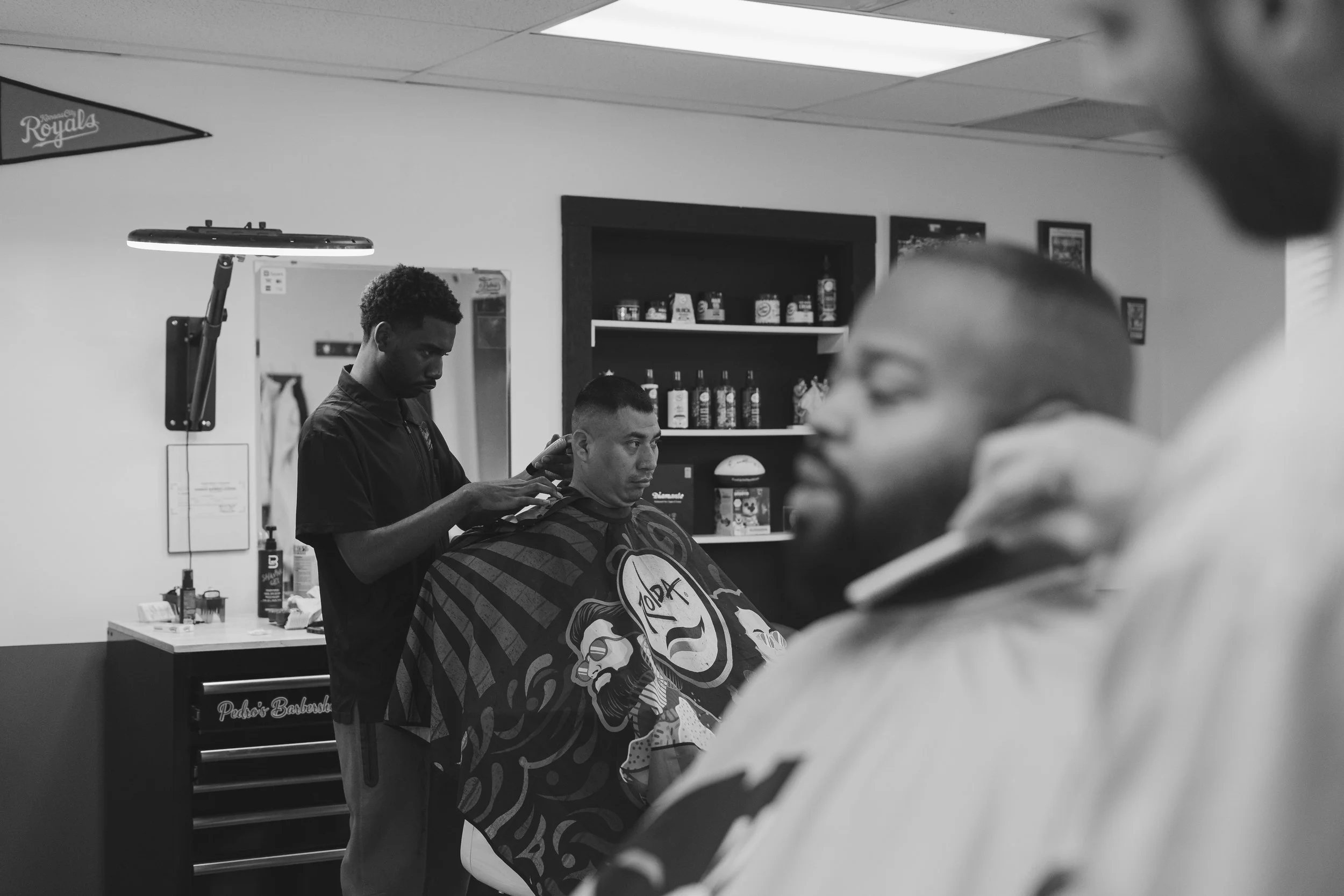 A person getting a haircut in a barbershop with two other clients seated. The barber is cutting hair while the other client looks ahead. The background shows shelves with products and a mirror.