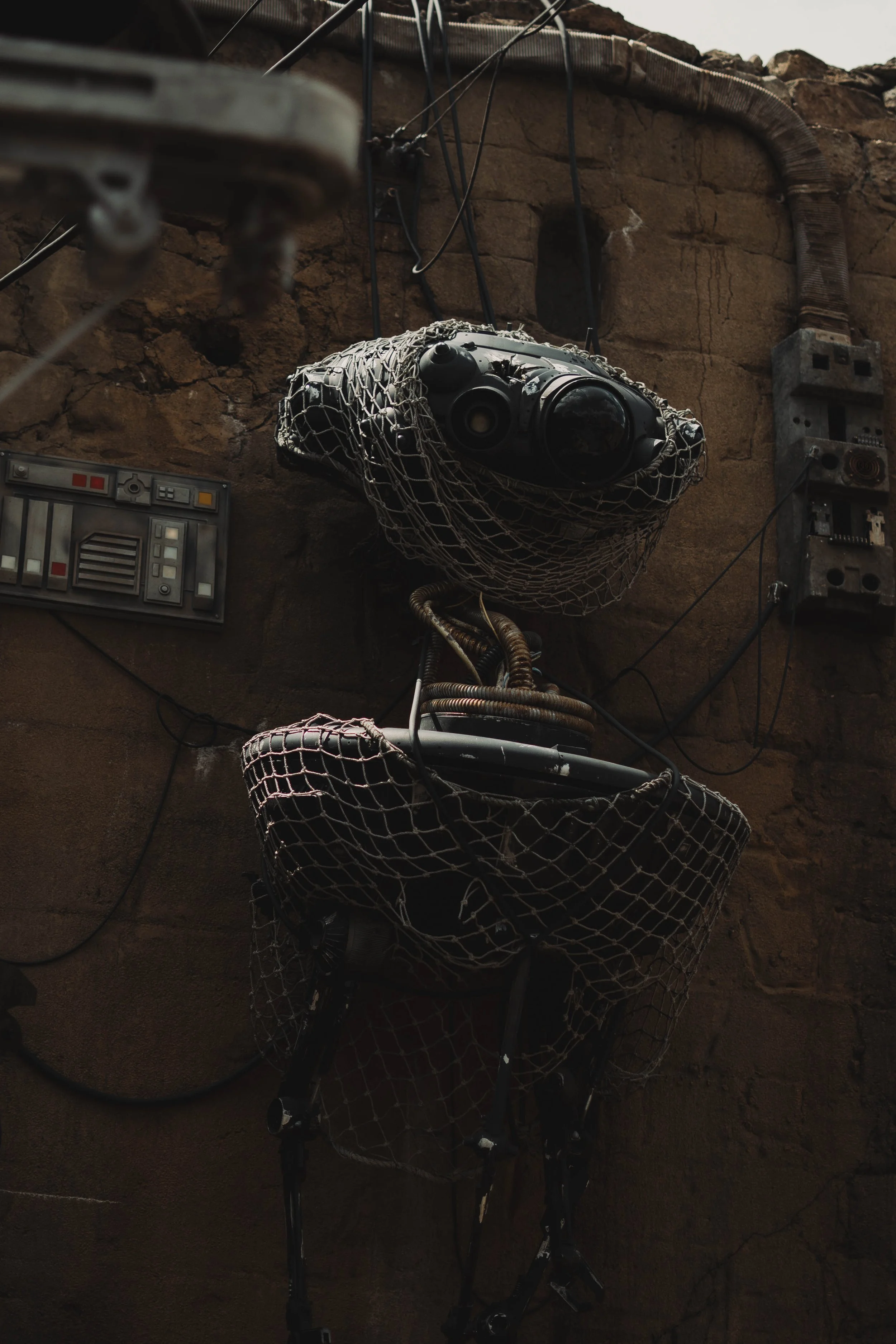 A robot designed to resemble a dog, covered with a cargo net, attached to a wall with various electrical cables and boxes.