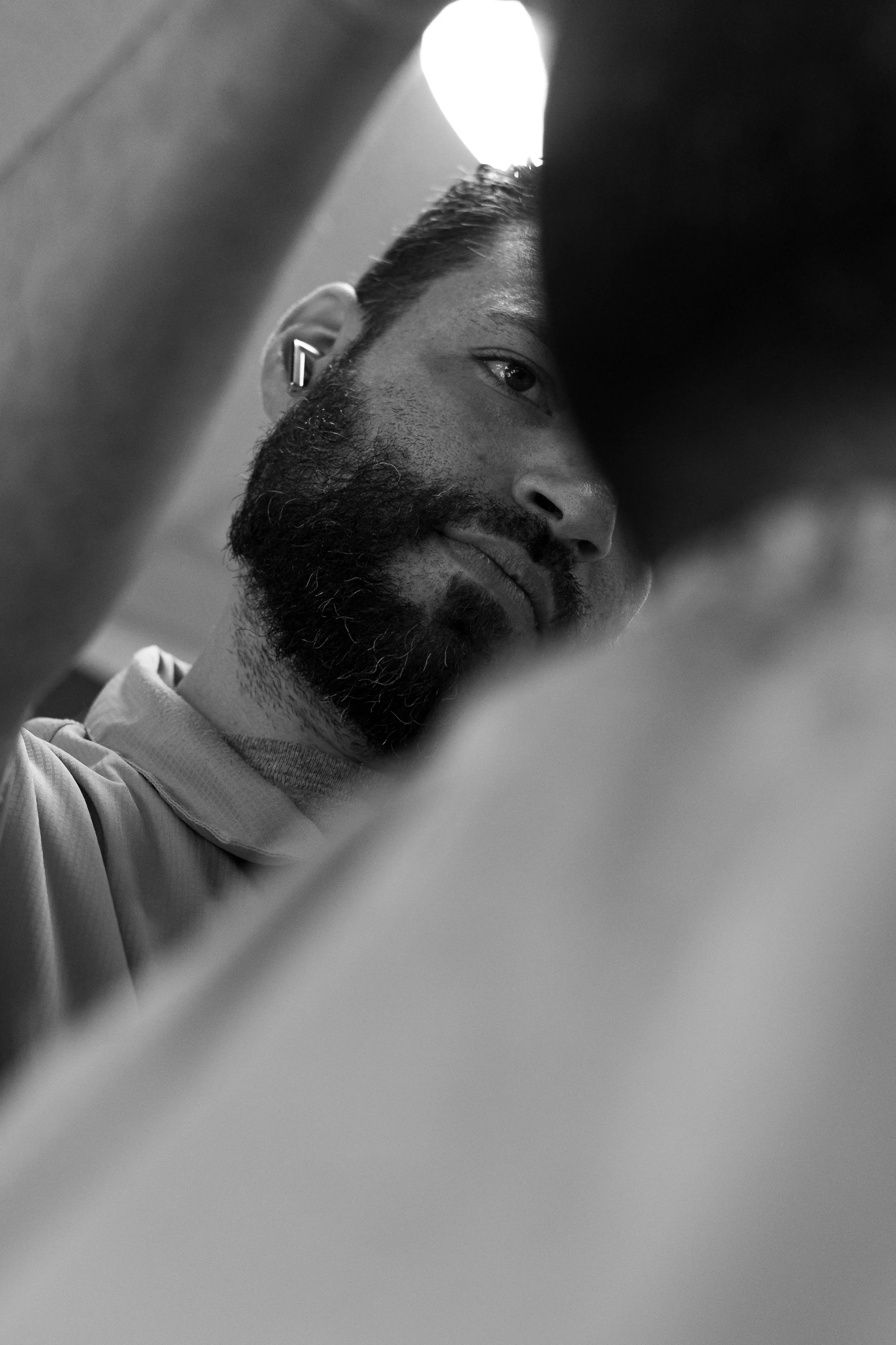 Close-up black and white photo of a man with a beard and short hair, looking down, seen through an opening with blurred foreground.
