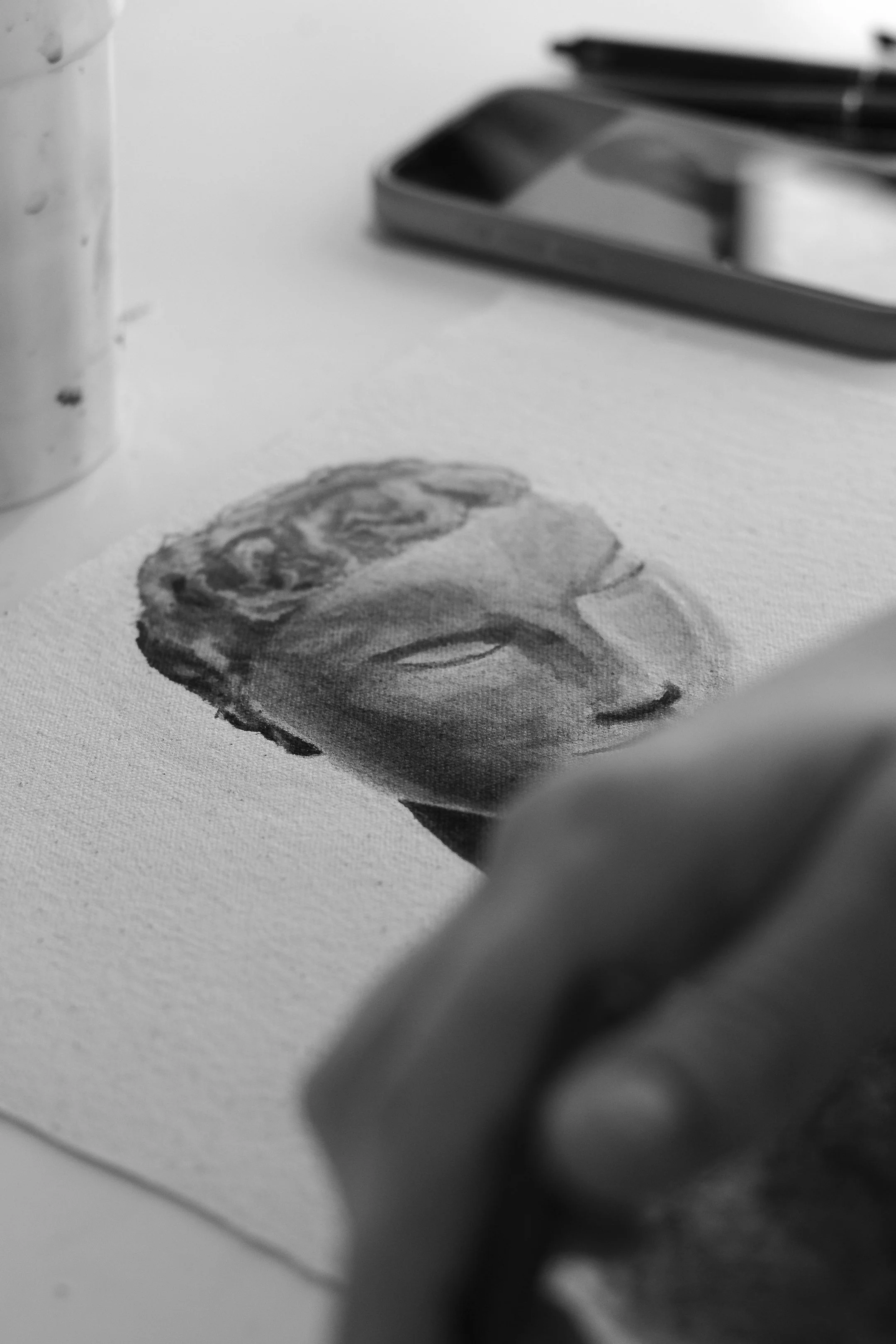 Black and white photograph of an artist's hand painting a portrait of a person's face on canvas, with art supplies in the background.