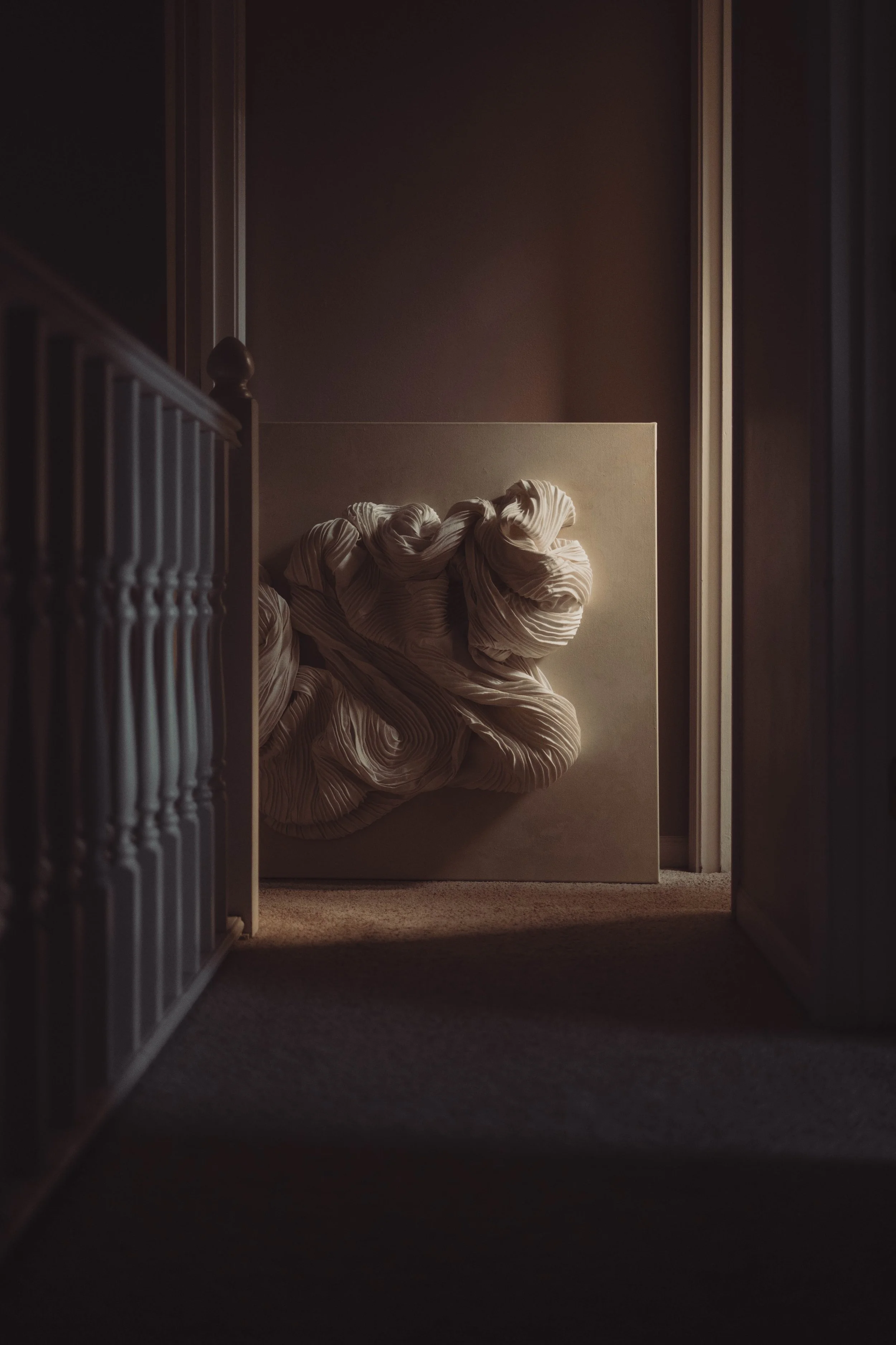 A framed, textured abstract artwork leaning against a wall in a dimly lit hallway with a carpeted floor.