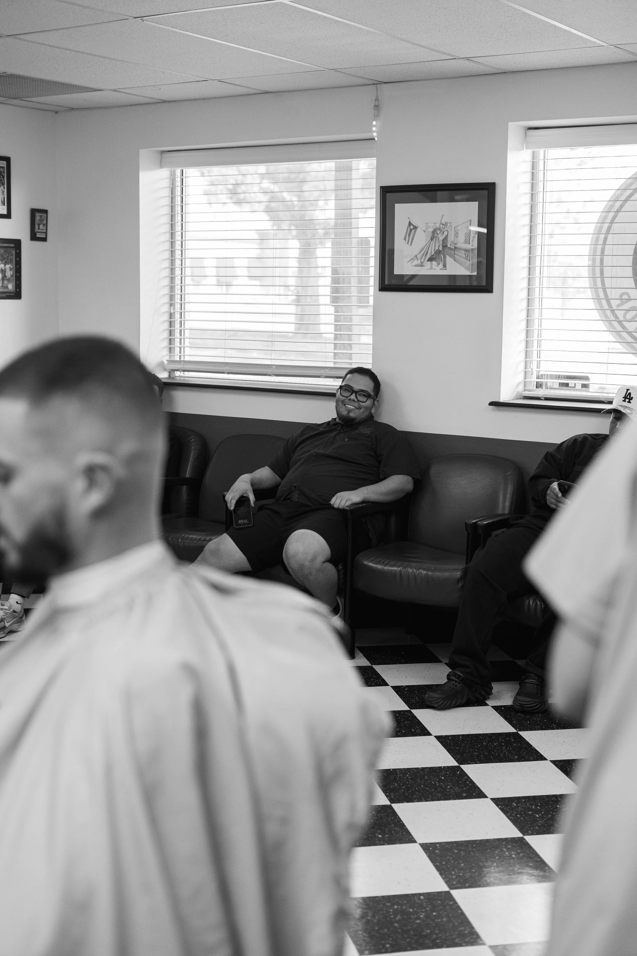 A group of men sit in a waiting room with black and white checkered floor, some seated on a black leather couch, one person smiling while holding a phone, and others visible in the foreground and background.