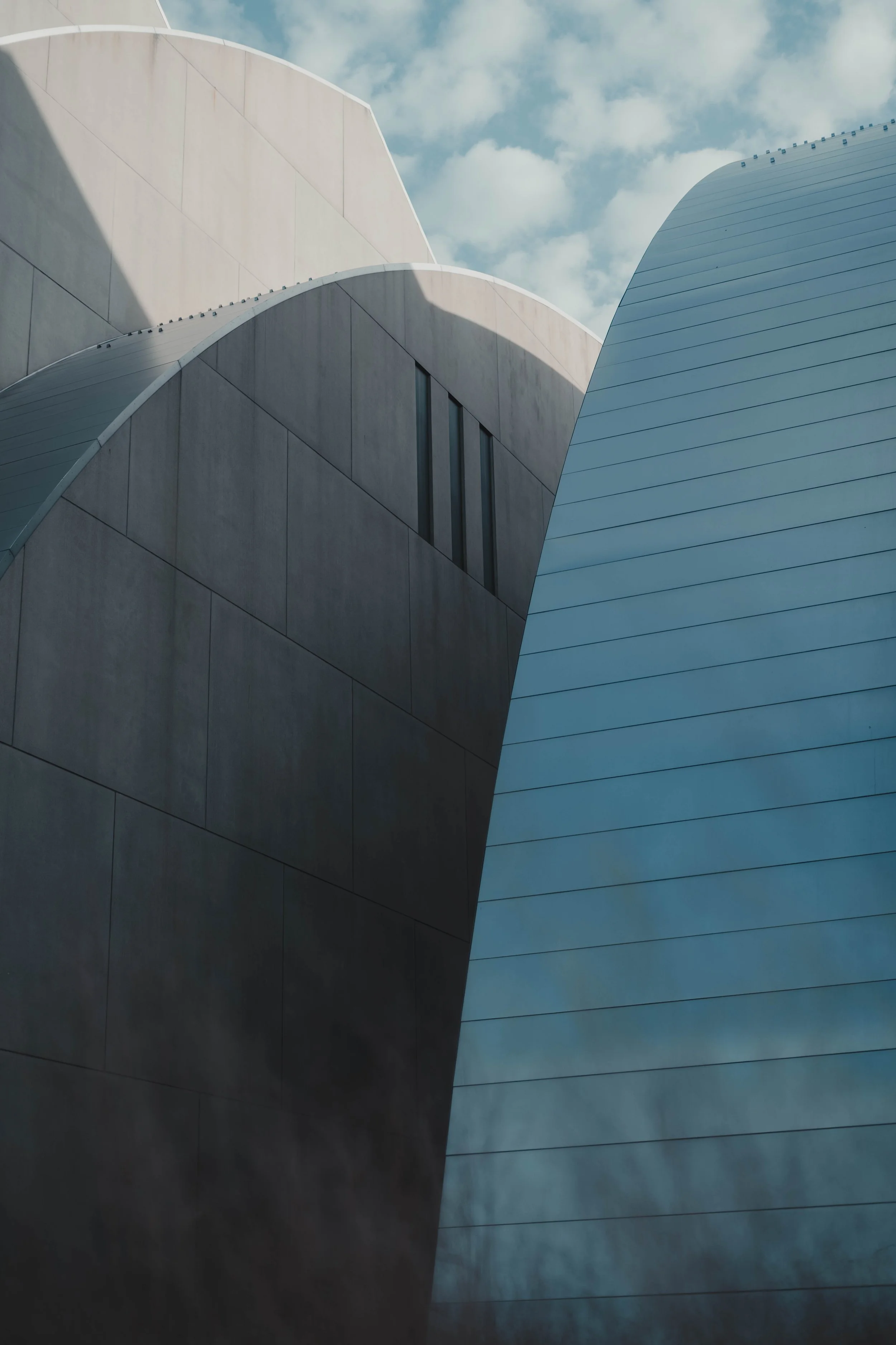 Close-up of modern architectural buildings with curved, metallic and concrete facades under a partly cloudy sky.