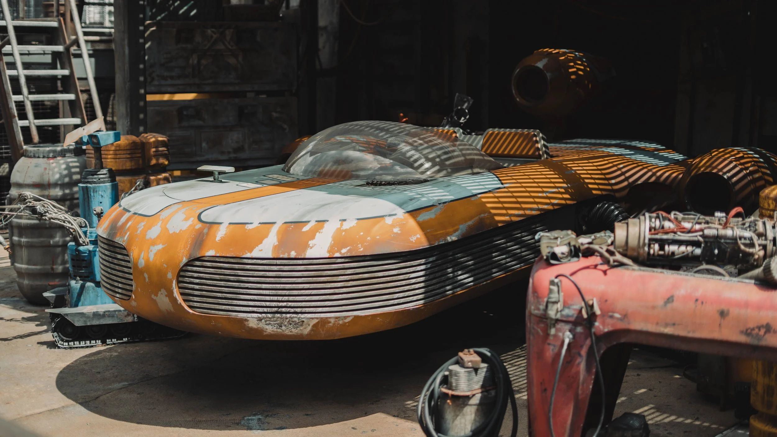 A vintage-style race car with a weathered orange and white paint job, stored in a workshop or garage surrounded by tools and equipment.