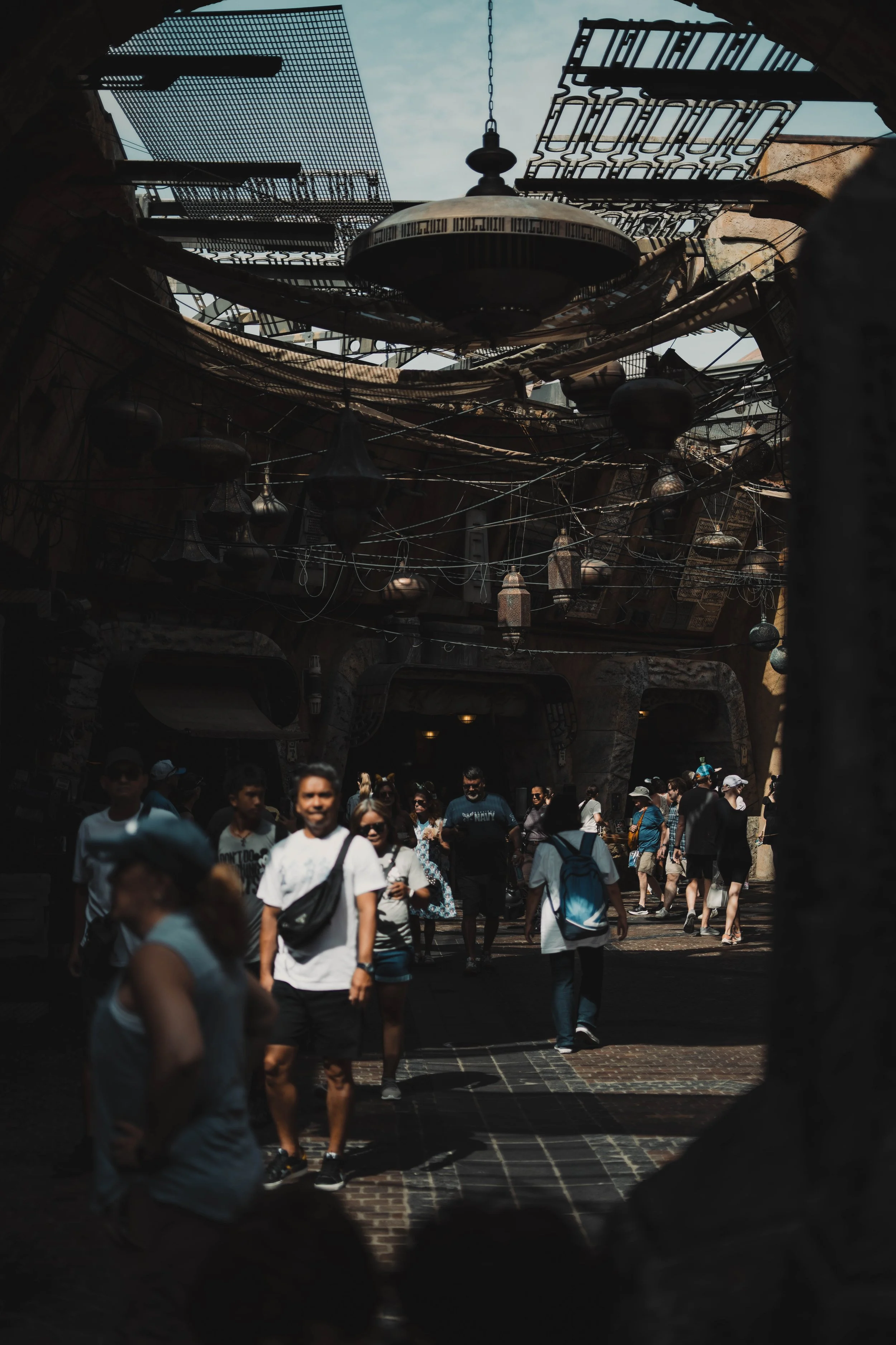 People walking through a shaded, bustling street with hanging lanterns and decorative metalwork overhead.