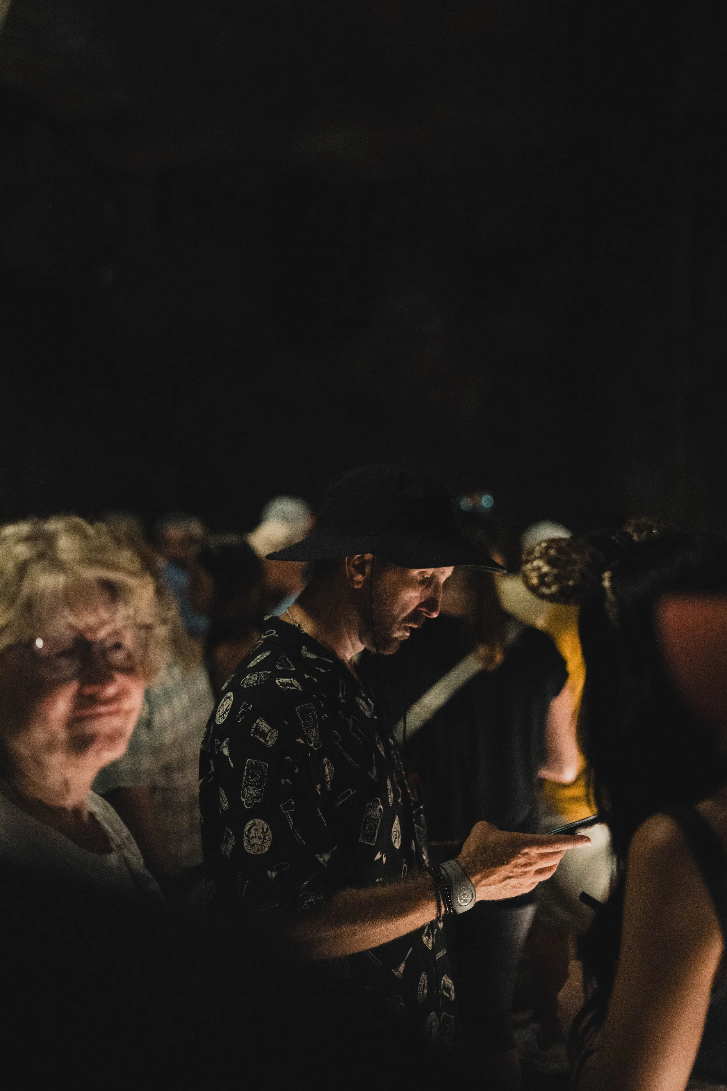 A man wearing a dark hat and a shirt with drinks and food prints looks at his phone in a dimly lit crowd.