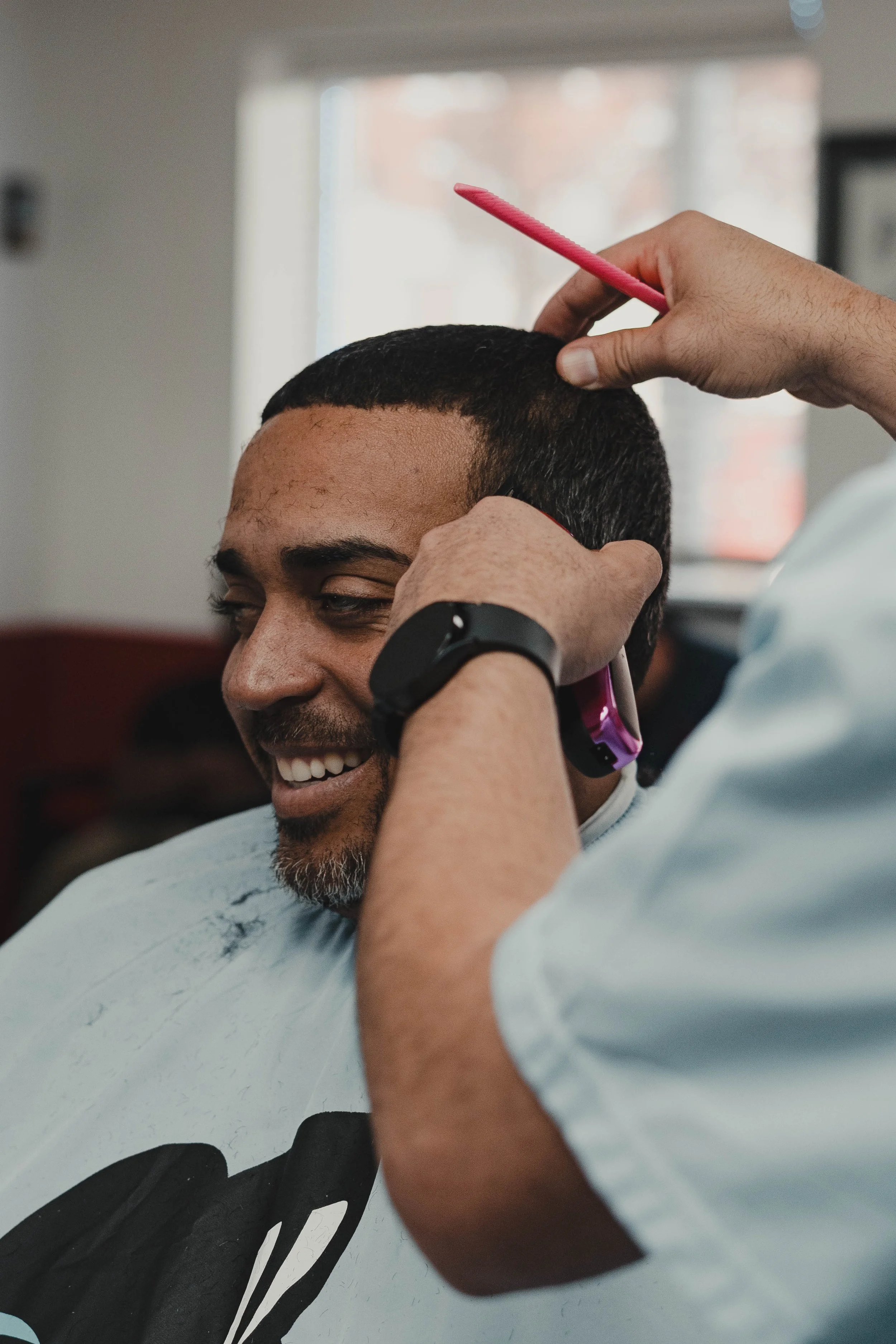 Man getting a haircut in a barbershop, smiling, with the barber using a pink comb and trimming his hair.
