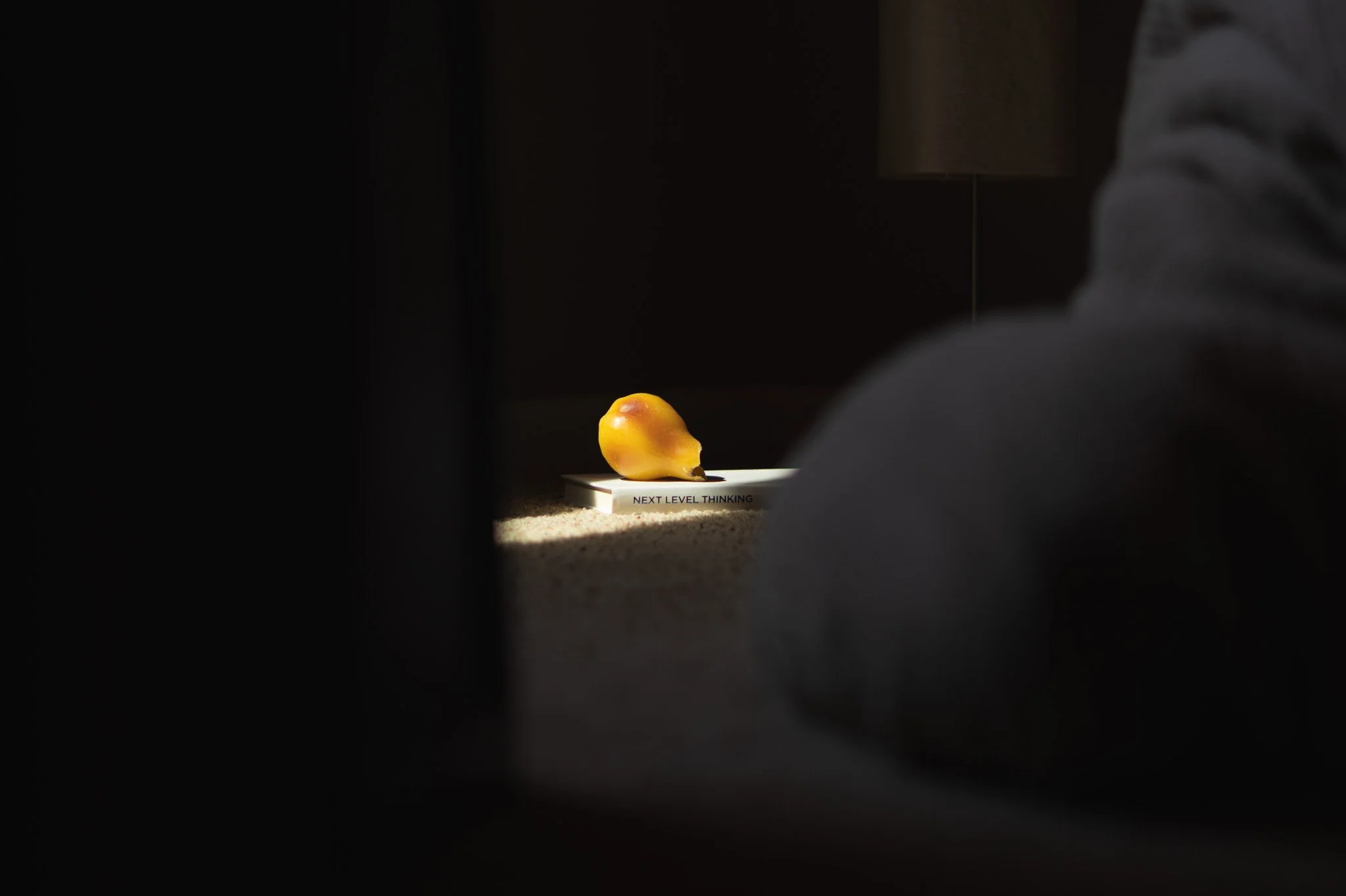 A yellow fruit, possibly a pear, resting on a white tray with a caption that reads 'NEXT LEVEL THINKING,' illuminated by a beam of light in a dark room, with a person sitting nearby.