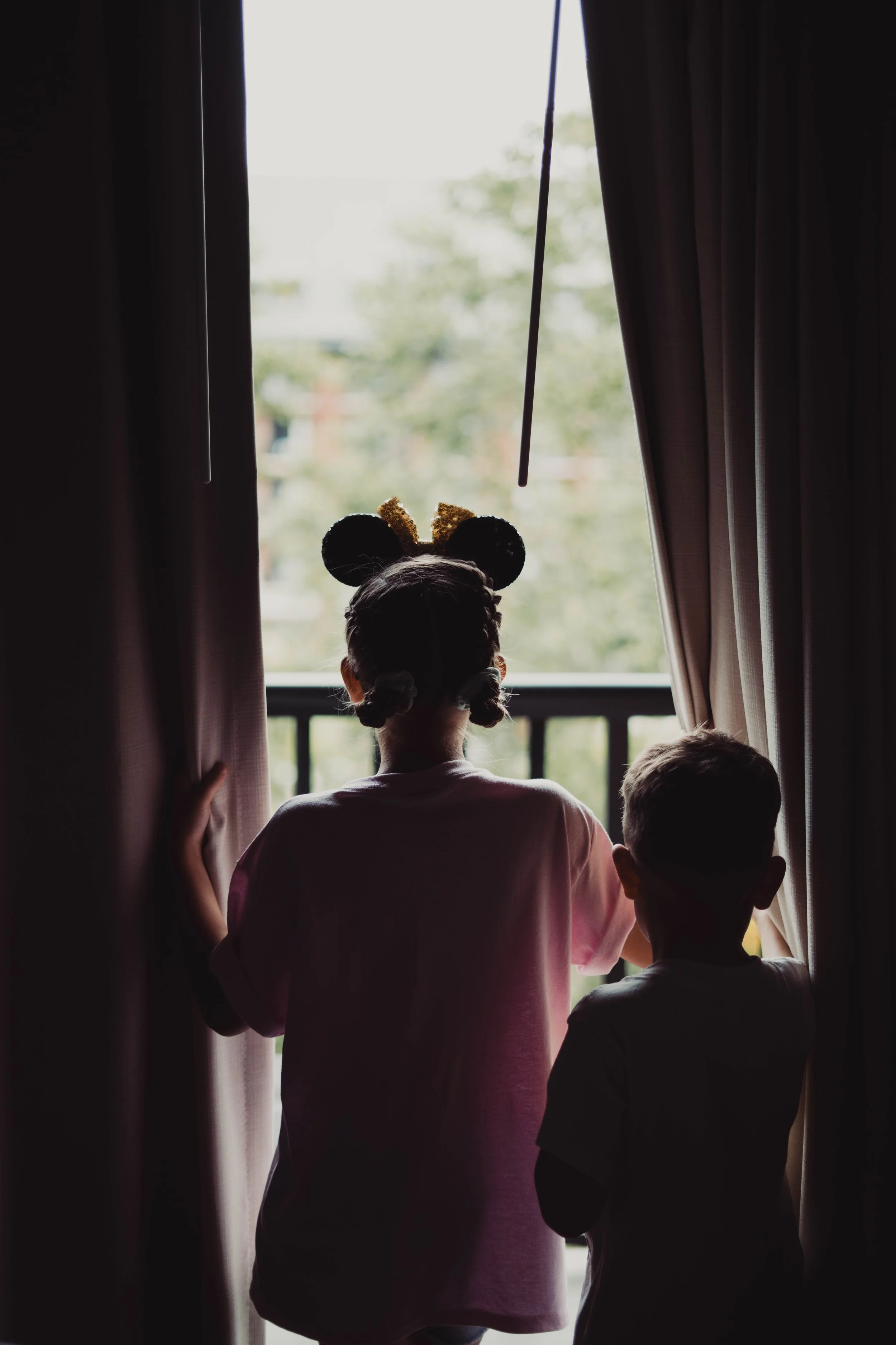 Two children, a girl with pigtails and a girl with Mickey Mouse ears, are standing near an open door looking outside, with curtains on either side.
