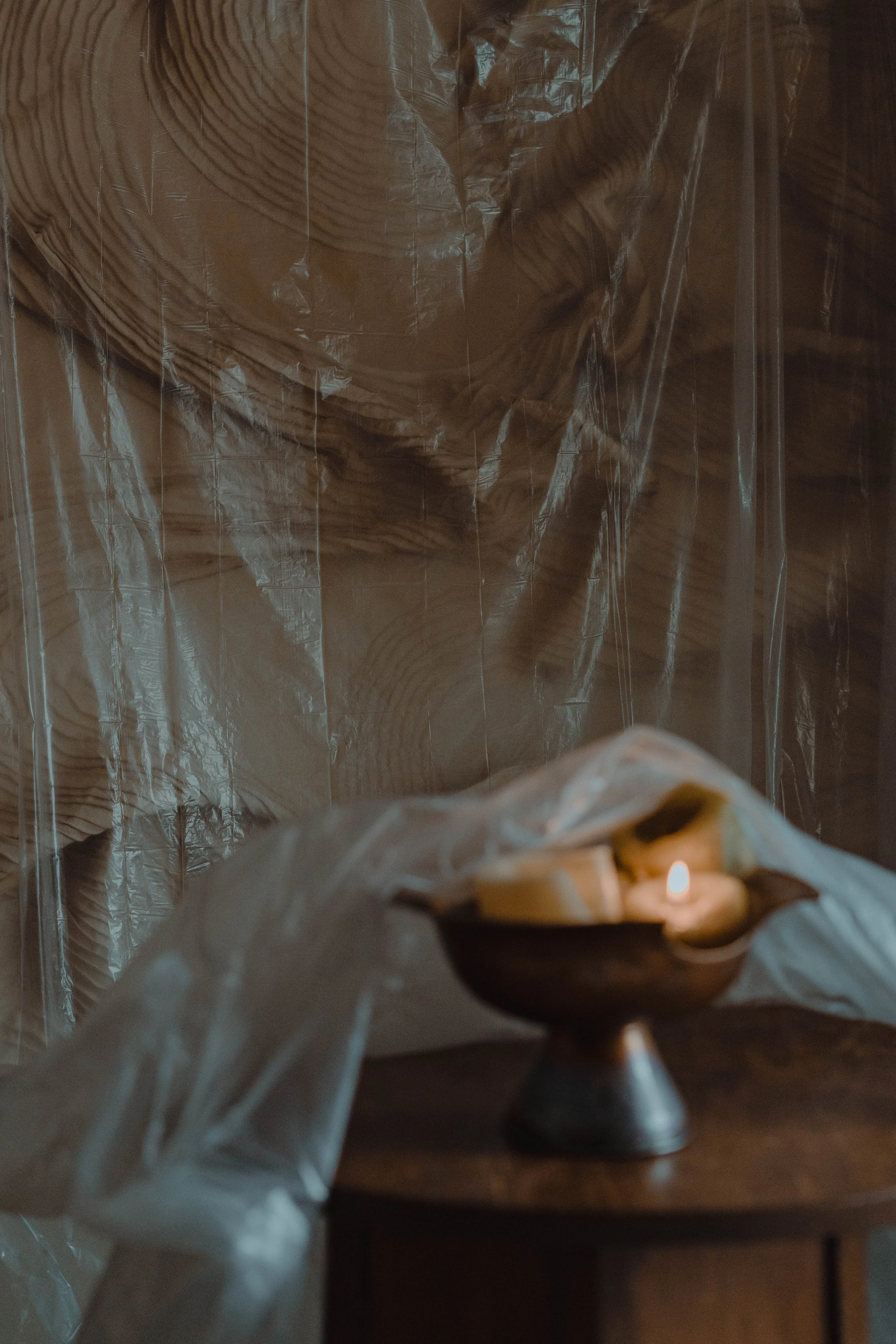 A small lit candle in a bowl on a wooden table, with a plastic sheet partially covering the bowl, and a textured, draped backdrop behind.