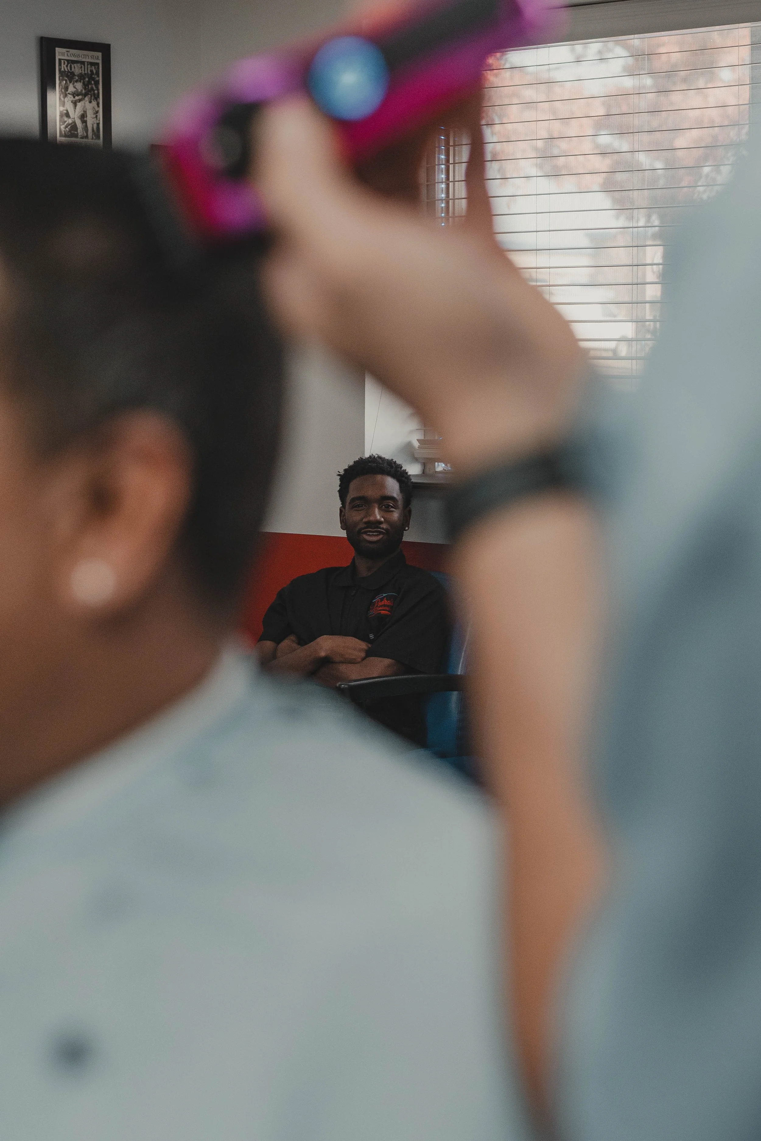 A man sitting with folded arms in a room with large window blinds, looking towards the camera, while a woman in the foreground is adjusting her pink sunglasses.