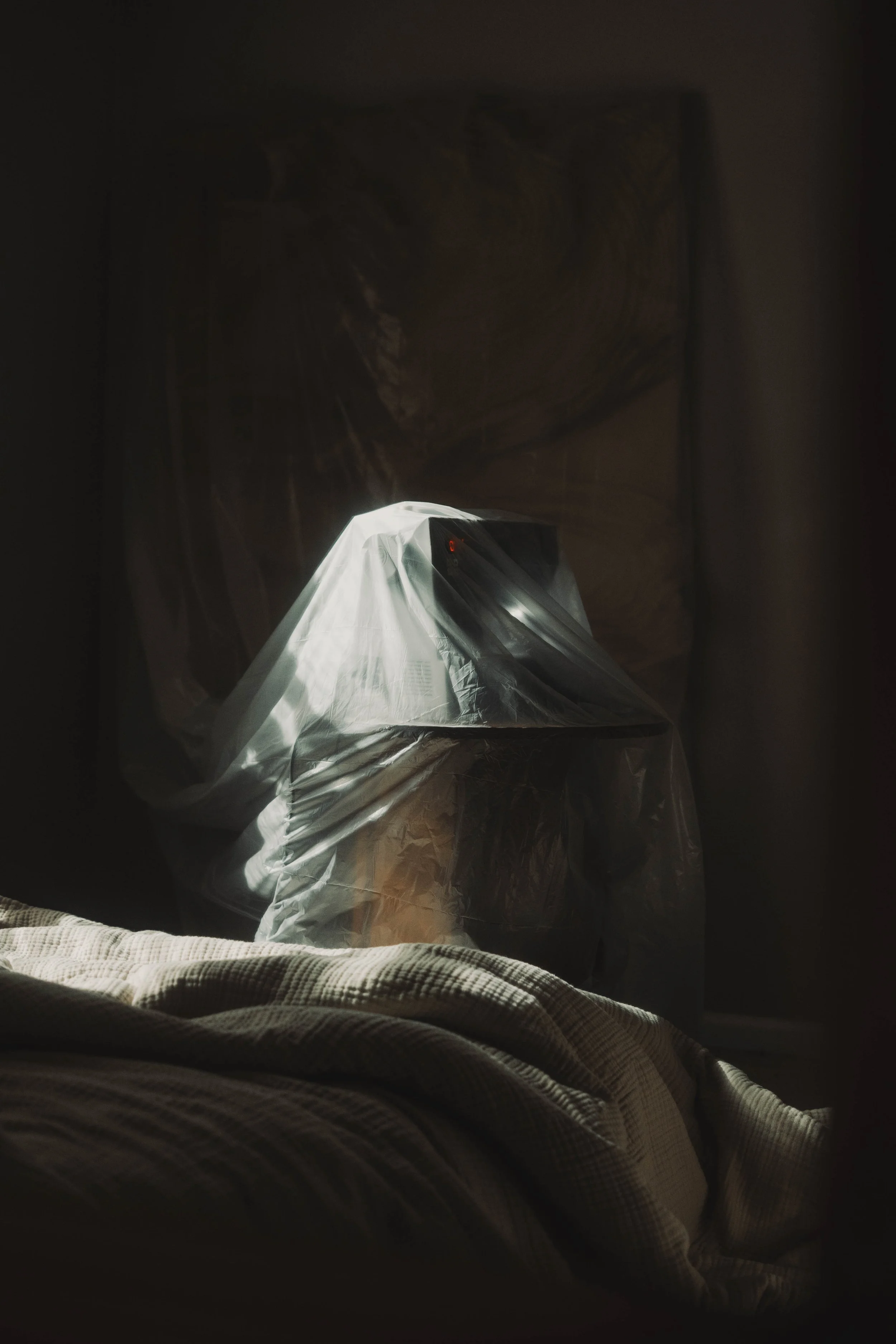 A chair covered with a plastic sheet in a dimly lit room with a bed in the foreground.