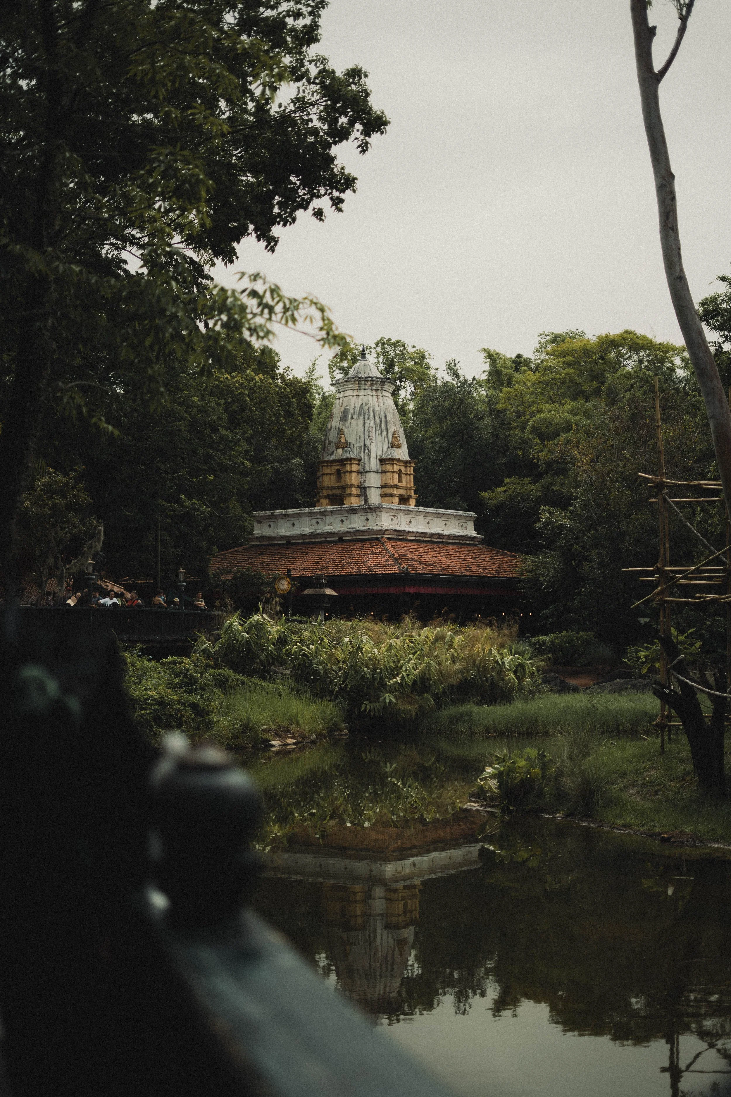 A temple or monument with a domed top situated among trees, with its reflection visible in a body of water in the foreground.