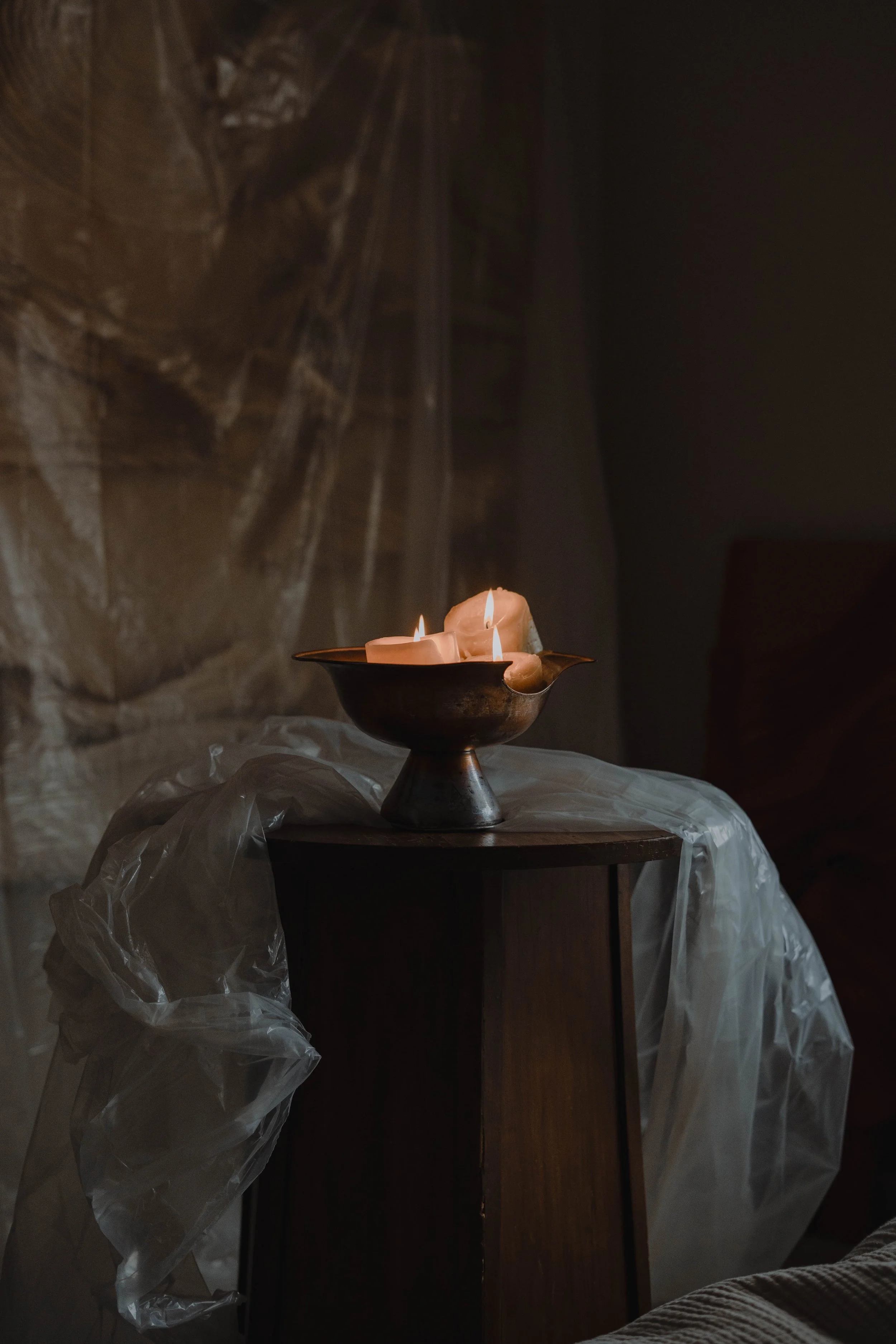 A metal bowl with three lit candles sits on a dark wooden stand, draped with translucent plastic. The background is dark with a faint, abstract artwork.