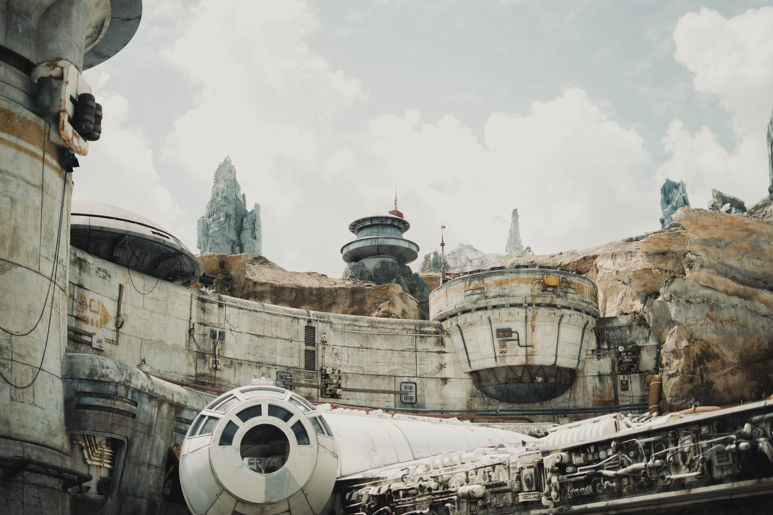 A futuristic, desert-like landscape with large, weathered industrial structures and rocky formations under a cloudy sky.