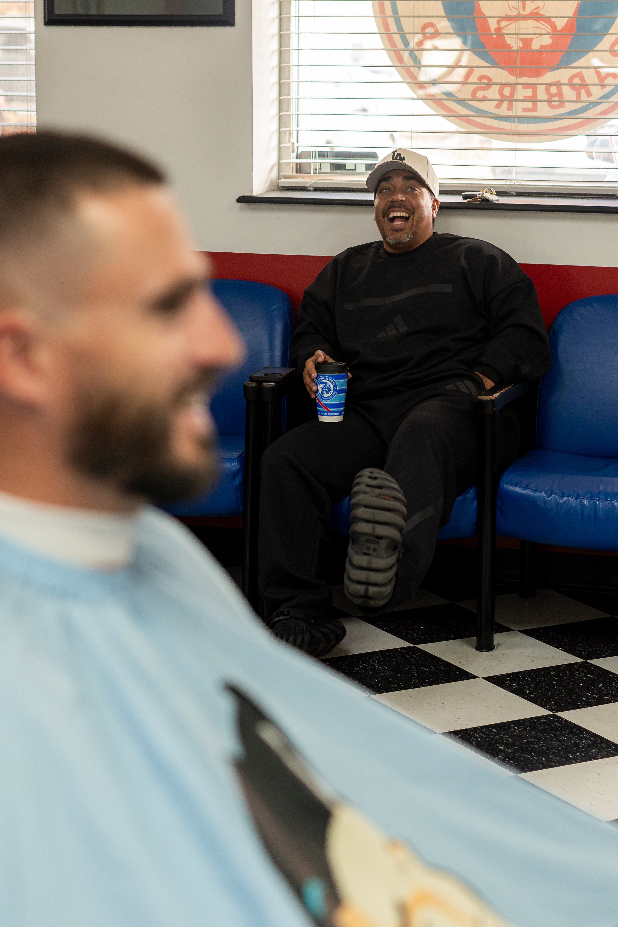 A man in a black sweater and light gray baseball cap sitting on a blue chair at a restaurant, laughing, holding a branded cup, with another man in disposable barber cape in the foreground.