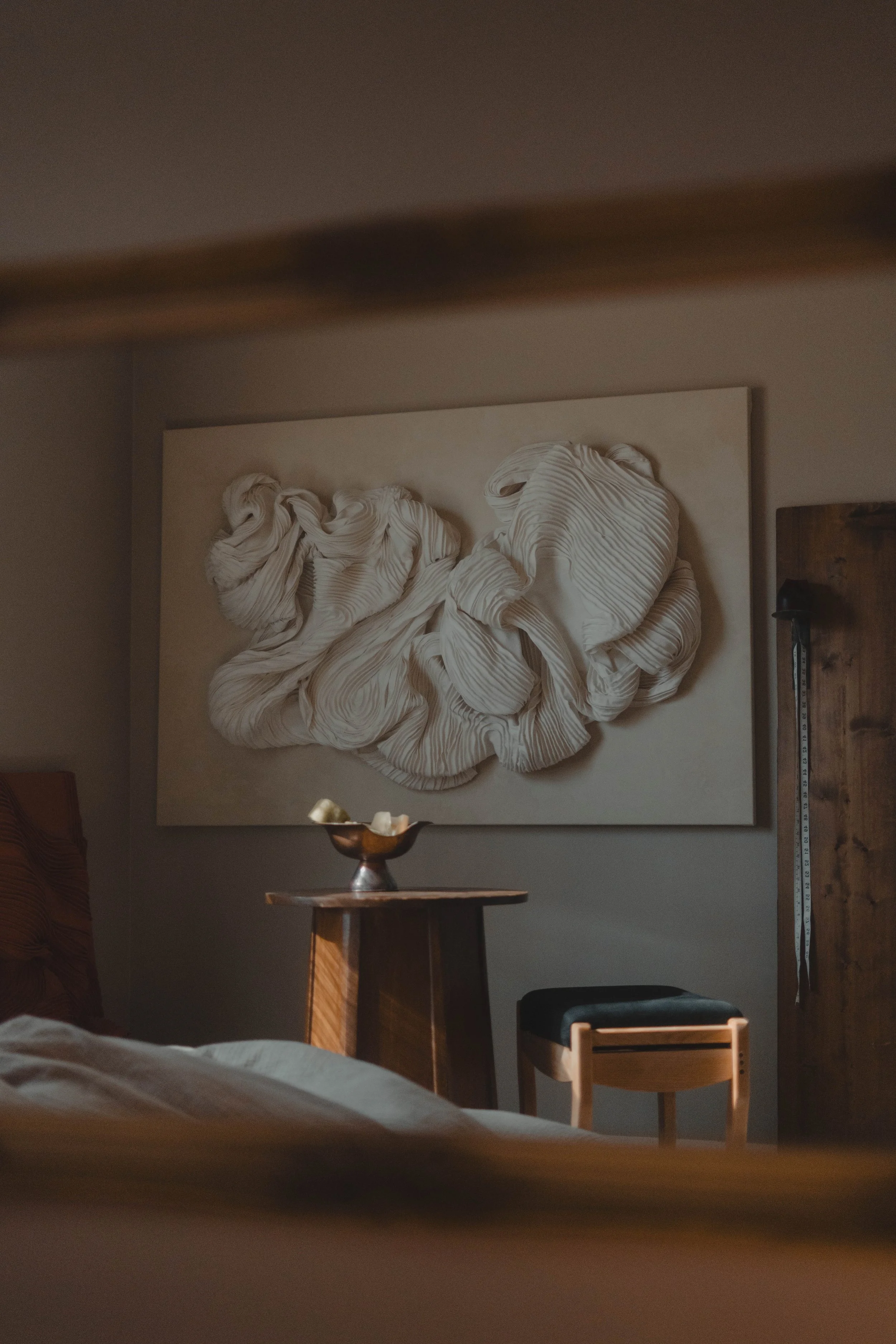 Interior of a bedroom featuring a textured white artwork on the wall, a wooden bedside table with a metallic bowl, and a black and wooden chair.