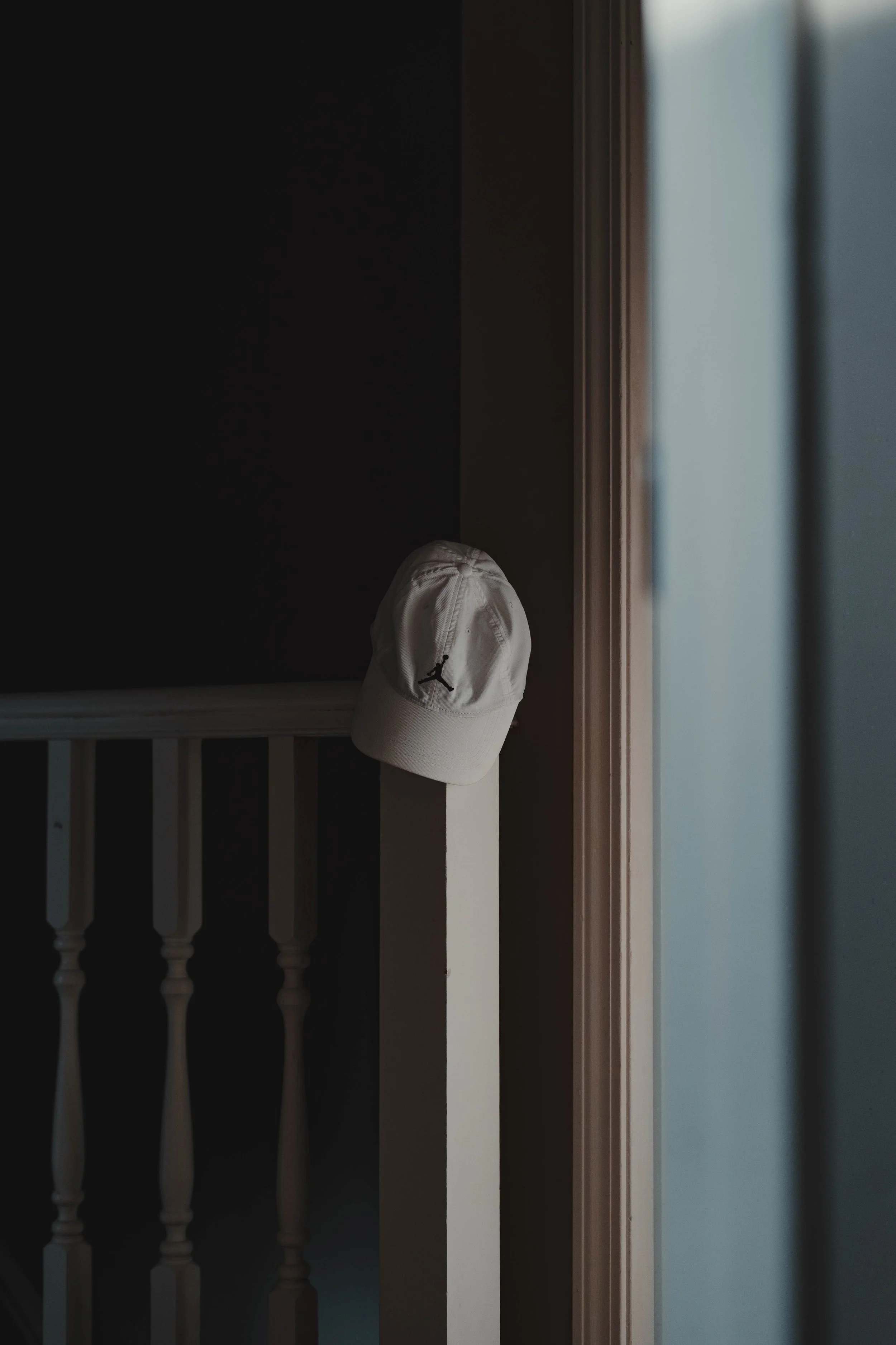 A white baseball cap with a black Jumpman logo hanging on a white feature near a staircase inside a house, with light coming in from a window.