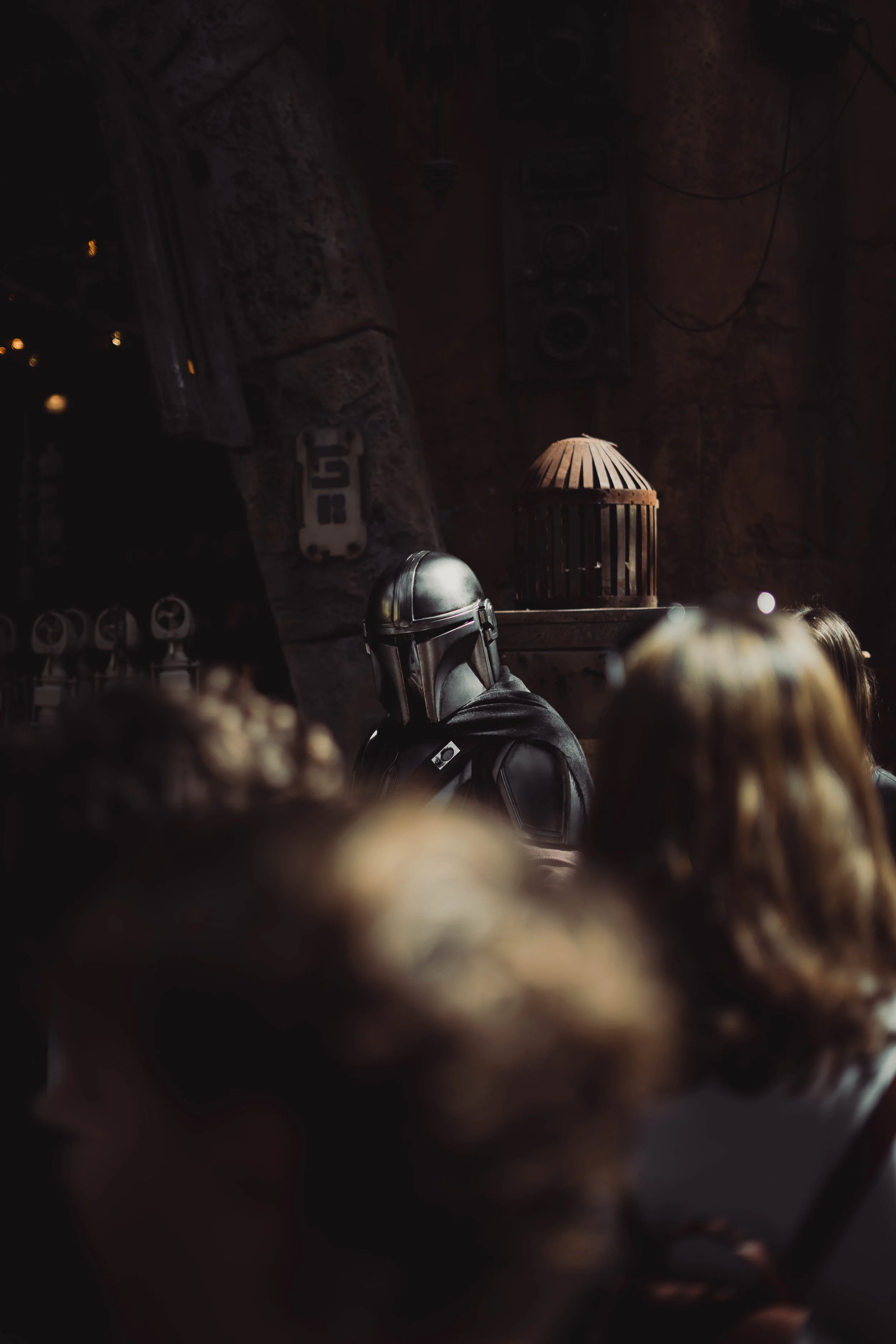 A person dressed as Boba Fett from Star Wars in a dark setting with people in the foreground and a cage-like structure behind.