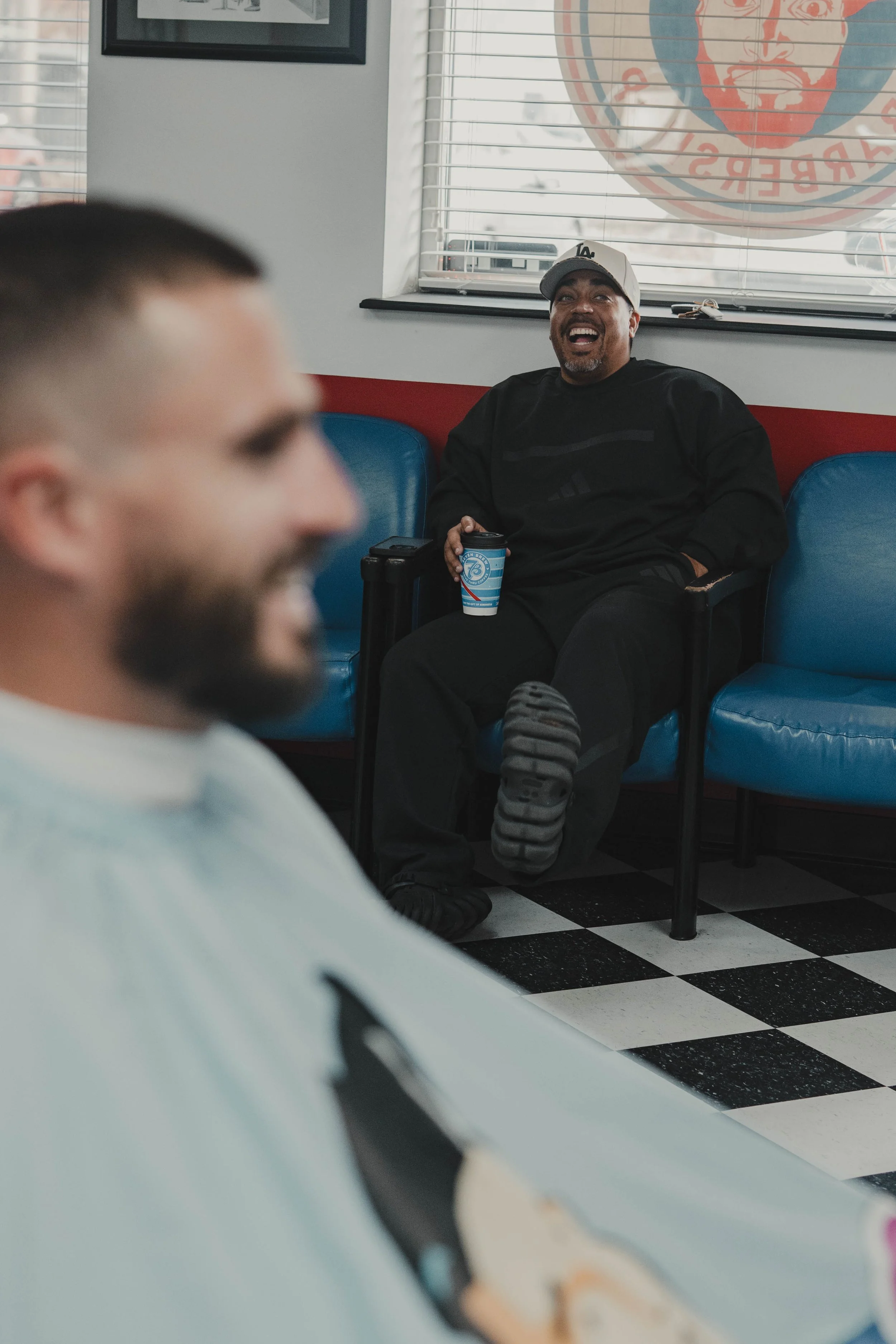 Two men in a retro-style diner, one in focus sitting on blue chairs, holding a coffee cup, and smiling, the other out of focus in the foreground.
