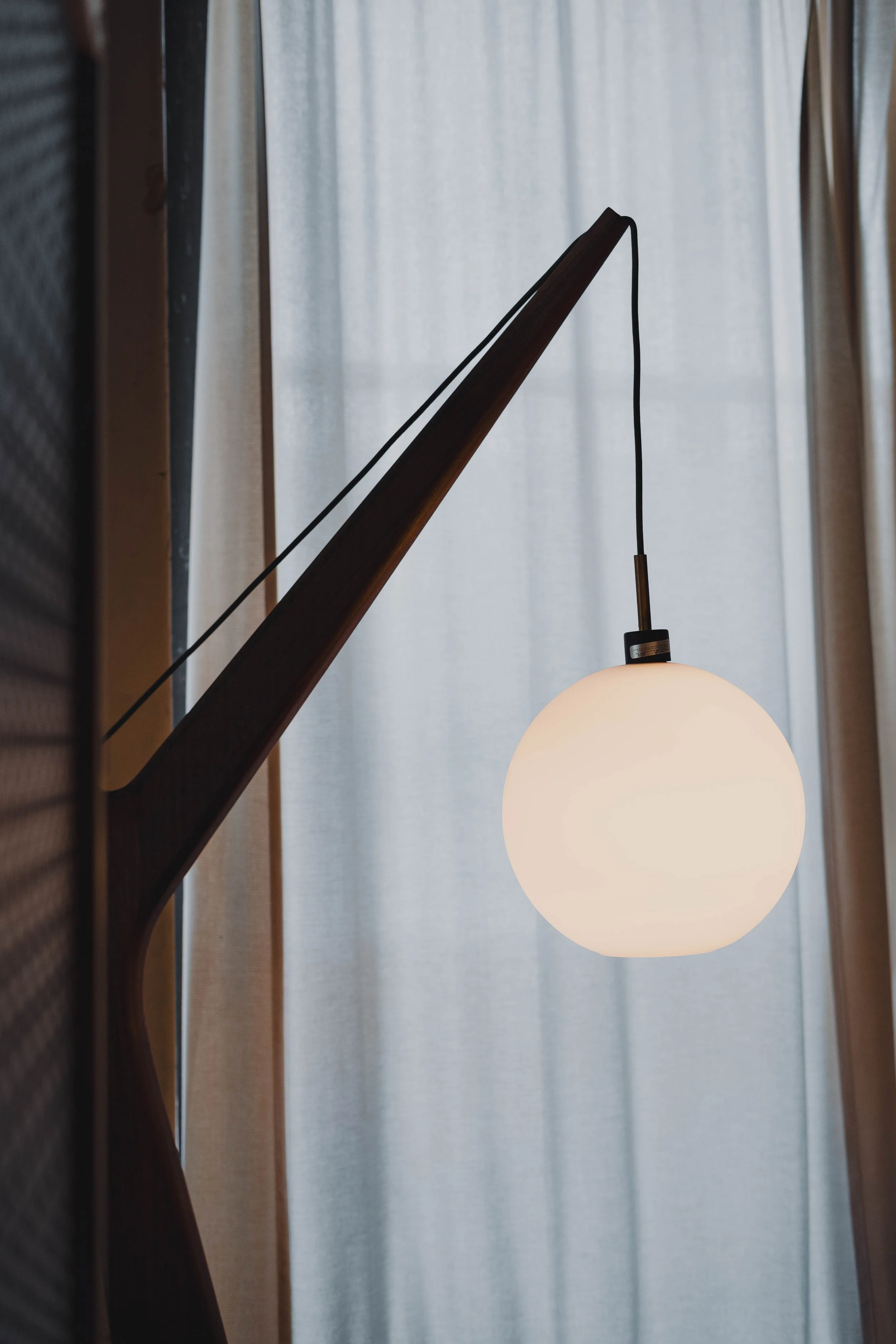 A modern hanging lamp with a spherical white shade and a dark wooden arm, positioned in front of light-colored curtains.