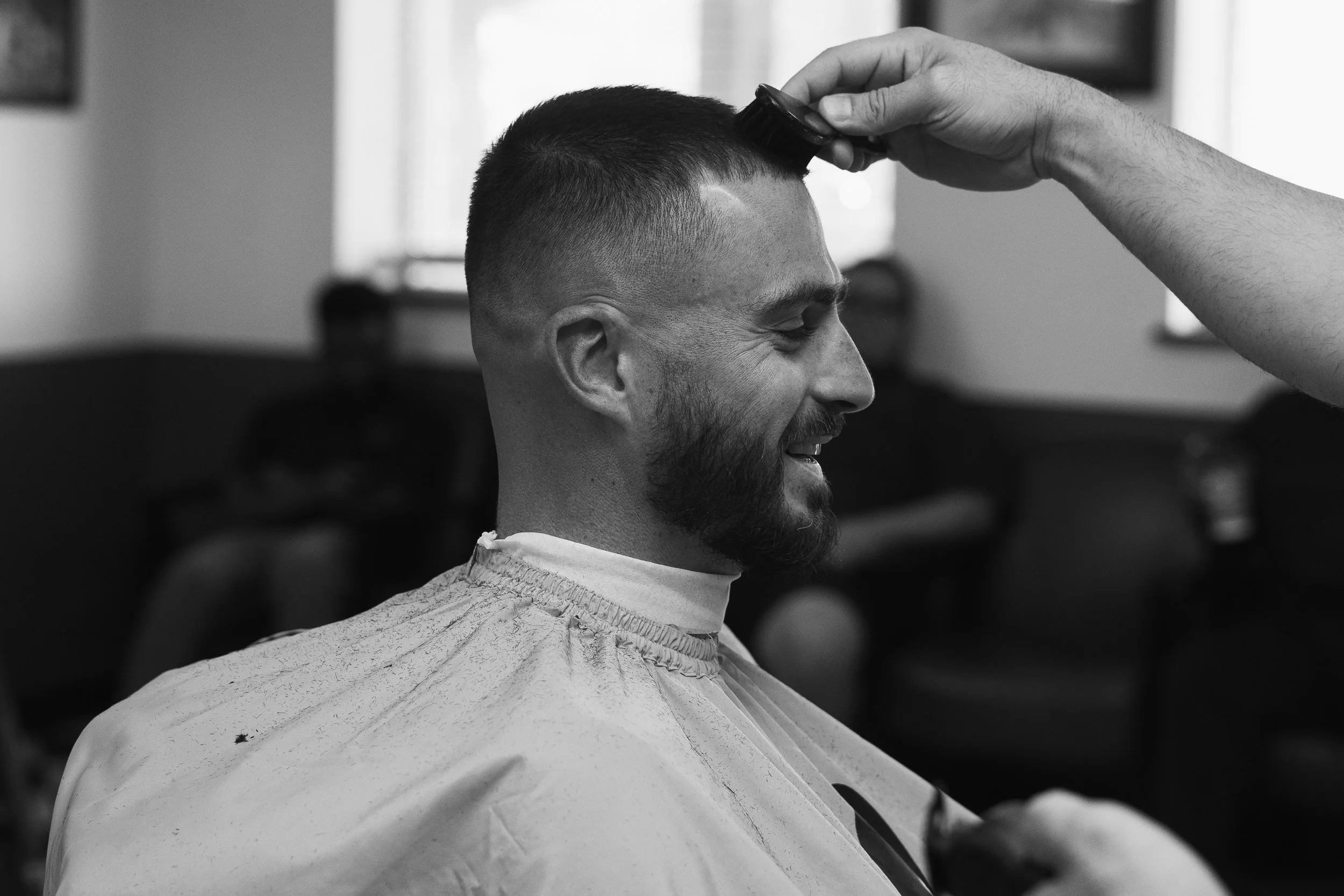 A man with a beard smiling as he gets a haircut in a salon, wearing a barber cap.